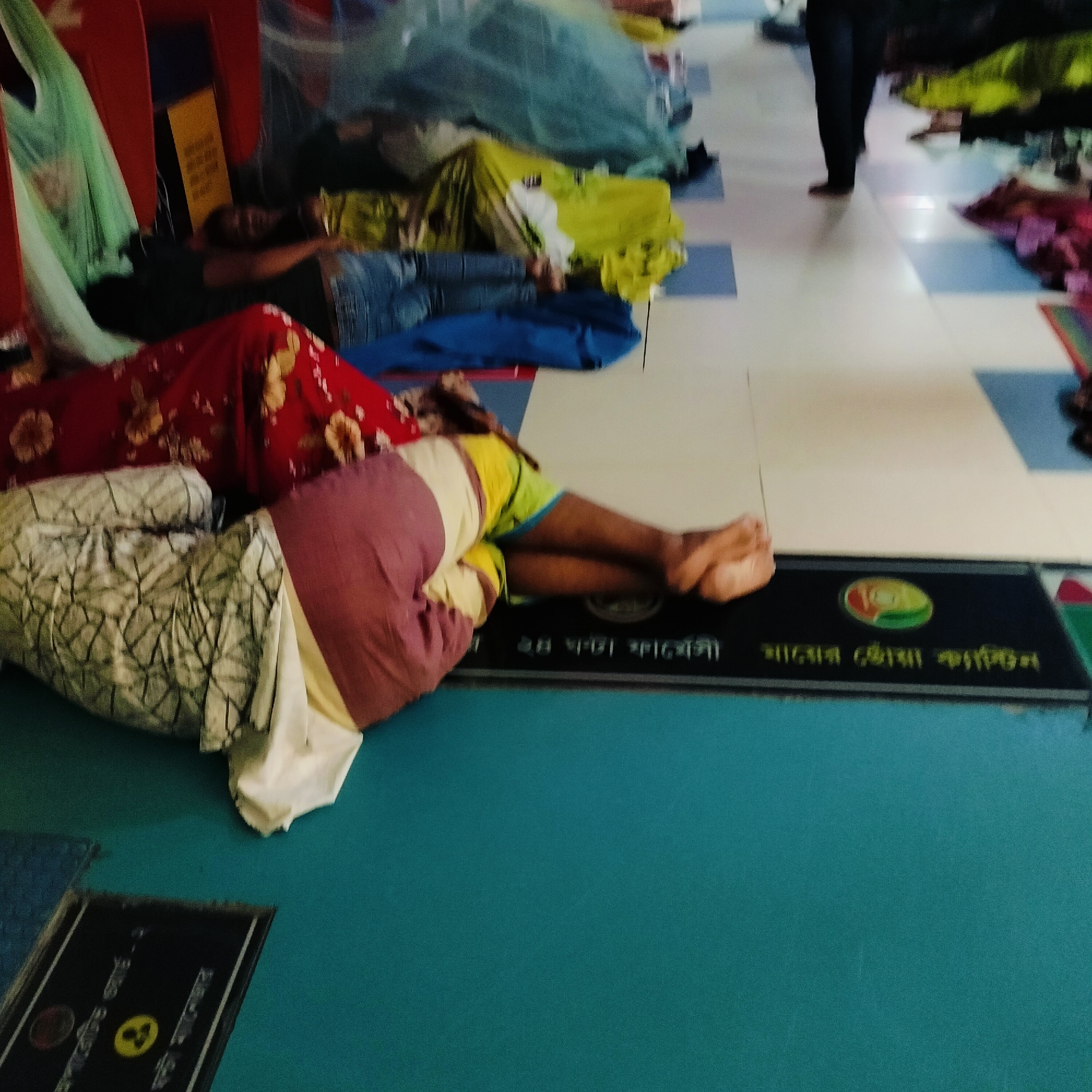 Several people are lying on the floor in what appears to be a public waiting area. The floor has alternating white and blue tiles. Some people are covered with blankets or clothing. In the background, a person is walking across the tiled floor. In the foreground, there is a small black mat with writing in what appears to be the Bengali language and a logo on it. Another sign, with a nuclear symbol, can be seen in the bottom-left corner of the frame. It is not possible to determine the exact location within Dhaka.