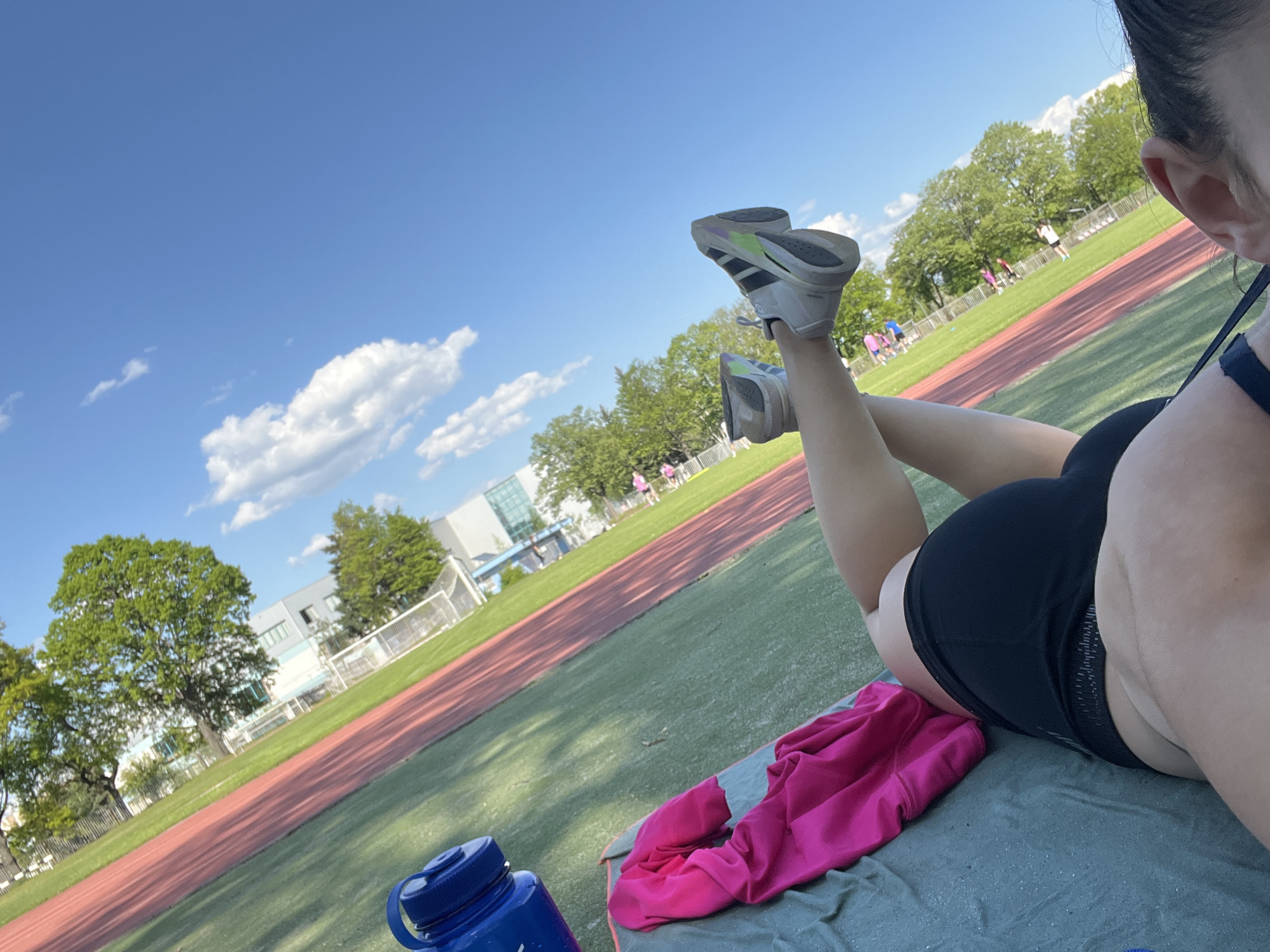 The image shows a person lying on a mat on a track, with their legs raised and feet wearing grey and white sneakers pointing towards the sky. A blue water bottle and a pink towel are placed near the mat. The track appears to be part of a sports complex with a red running track, a green field, a building in the background, and trees. Other individuals can be seen in the distance on the track. The sky is blue with scattered white clouds. The location is characterized by a track and field setting in the city of Unknown, Unknown.
