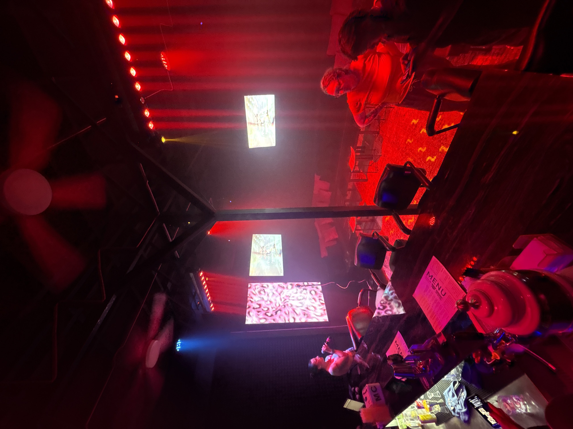 A dimly lit bar interior in Baan Bang Rak, Thailand, is illuminated primarily by red lighting. Three video screens hang from the ceiling, displaying abstract patterns and imagery. A man sits at the bar, holding a microphone and speaking. A menu and various objects, including bottles and glasses, are on the bar top. A ceiling fan rotates in the upper left of the frame.