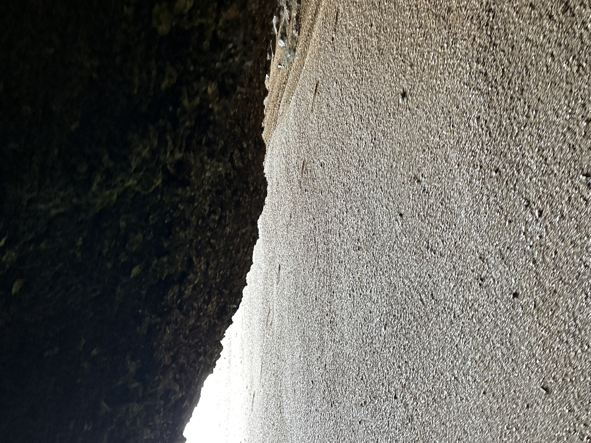 This is a ground-level, wide-angle shot taken from within a cave or overhang, looking out onto a beach. The foreground is dominated by the rough, dark texture of the overhang, which obscures the upper portion of the image. Below the overhang, the beach is covered in small, light-colored pebbles and sand, with a faint sheen suggesting moisture. The light source appears to be behind the beach, creating a bright background and casting shadows from the pebble texture. There are no people or man-made objects visible in the scene, and the environment appears to be natural. The specific location in Krasom, Thailand, cannot be determined from the visual information provided.