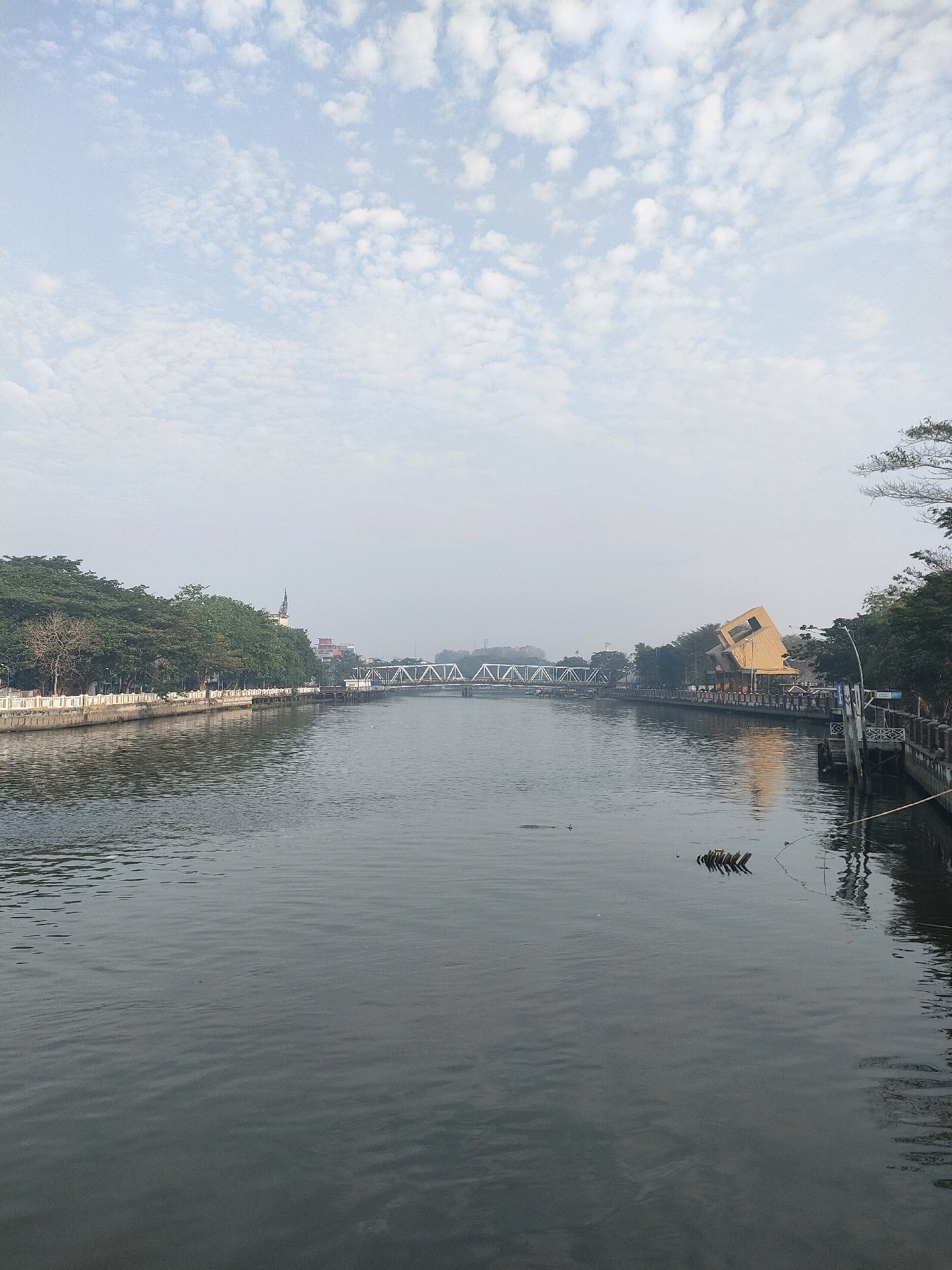 The image depicts the Martapura River in Banjarmasin, Indonesia, under a cloudy sky. A white steel truss bridge spans the river in the distance. On the left bank, a row of trees lines a promenade with a white railing. On the right bank, there is a distinctive orange building with a slanted roof, situated along the waterfront. The river itself is calm with ripples reflecting the muted sky. Several wooden posts are visible jutting out of the water near the right bank. The scene appears to be an urban waterfront area, likely a public space for recreation or transit.