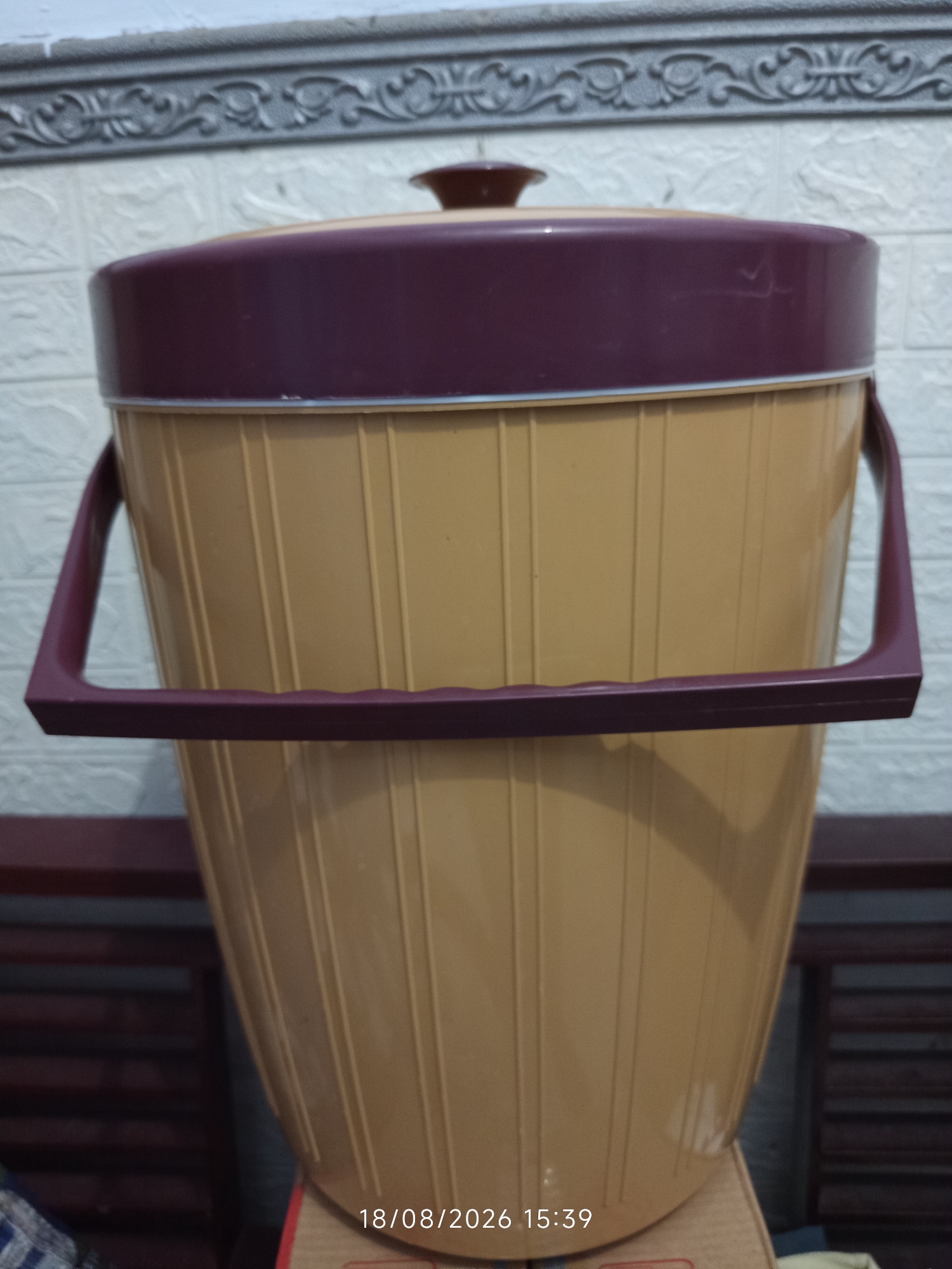 An indoor, eye-level, medium shot shows a large, tan-colored, plastic beverage dispenser with a dark plum-colored lid and handles, positioned on top of a cardboard box. The dispenser has vertical ridges and a handle on each side, running horizontally across the lower portion of the dispenser. The lid is slightly domed with a smaller, darker plum-colored knob on top. In the background, a white tiled wall is visible with a decorative gray border running horizontally across the top. The lower portion of the background consists of the dark brown slats of furniture. A timestamp in the bottom right corner reads "18/08/2026 15:39". There are no people or animals visible in the image. The location is identified as Canggu, Indonesia, based on the provided context.