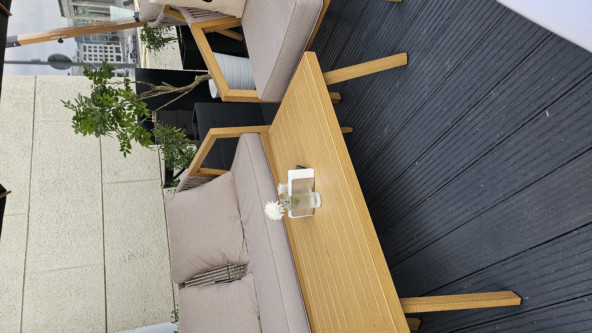 The image shows an outdoor seating area with wooden furniture and a light-colored stucco wall. A light brown slatted coffee table is in the foreground, with a small glass vase containing a white flower and a small stack of white napkins on it. Behind the table, a light brown sofa with matching cushions is visible, and to the right, a matching armchair. Two black planters with green plants are placed behind the sofa and armchair. The flooring is dark gray composite decking. In the background, through a window or opening, city buildings and vehicles can be seen, suggesting an urban setting. The overall impression is a modern, clean outdoor seating space that could be part of a cafe, restaurant, or residential patio.
