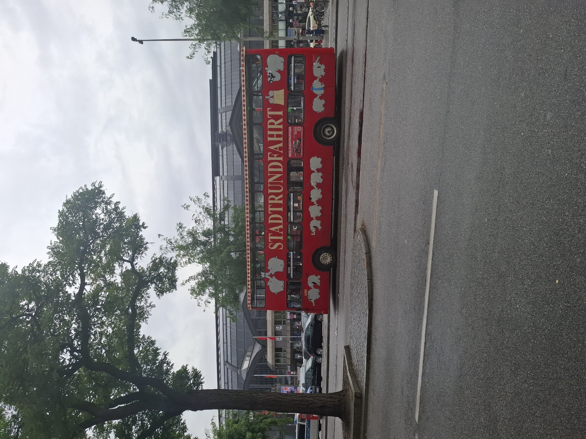 The image shows a red double-decker bus with the text "STADTRUNDFAHRT" (city tour) printed on its side, parked on a street in Hamburg, Germany. The bus is adorned with cartoonish illustrations of elephants. In the background, a large train station with a glass roof is visible, likely Hamburg Hauptbahnhof, the city's main railway station. Several cars and trees are also present in the scene, and the sky is overcast.
