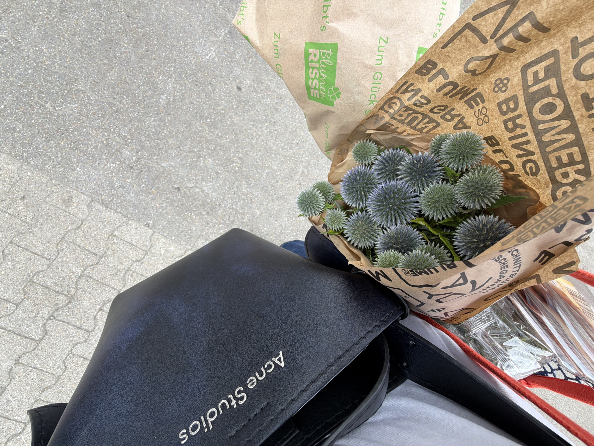 The image is a medium-angle, eye-level shot taken outdoors on a paved surface, likely a sidewalk or plaza. In the foreground, a bouquet of spiky, blue-green thistle-like flowers is wrapped in brown paper. The paper has a repeating pattern of text and graphics, including the brand name "BLUMEN RISSE" in green and "Zum Glück" in green. The text is partially obscured and some of it is upside down. To the left of the flowers is a black leather bag with "Acne Studios" embossed in silver on its surface. The bag appears to be a tote or shoulder bag. White and red fabric, along with a clear plastic-wrapped item, are visible beneath the bag. The background shows a light grey, textured pavement with visible seams between the paving stones. The overall impression is that of someone carrying shopping, including a floral purchase.