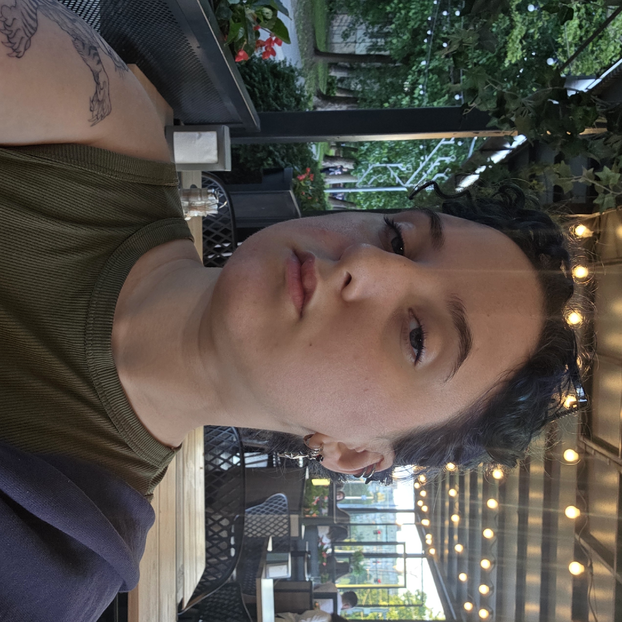 A medium shot captures a young woman sitting at a table on an outdoor patio, looking directly at the camera with a neutral expression. She has dark, curly hair styled in a bun and is wearing a green tank top. She has several earrings in her left ear, including a dangling star. A tattoo of a tiger is visible on her left arm. The background shows a covered outdoor seating area with string lights overhead, tables and chairs, and greenery. In the distance, through glass windows, a city scene with buildings and traffic lights is visible. The woman is seated at a wooden table with a napkin dispenser and cutlery.