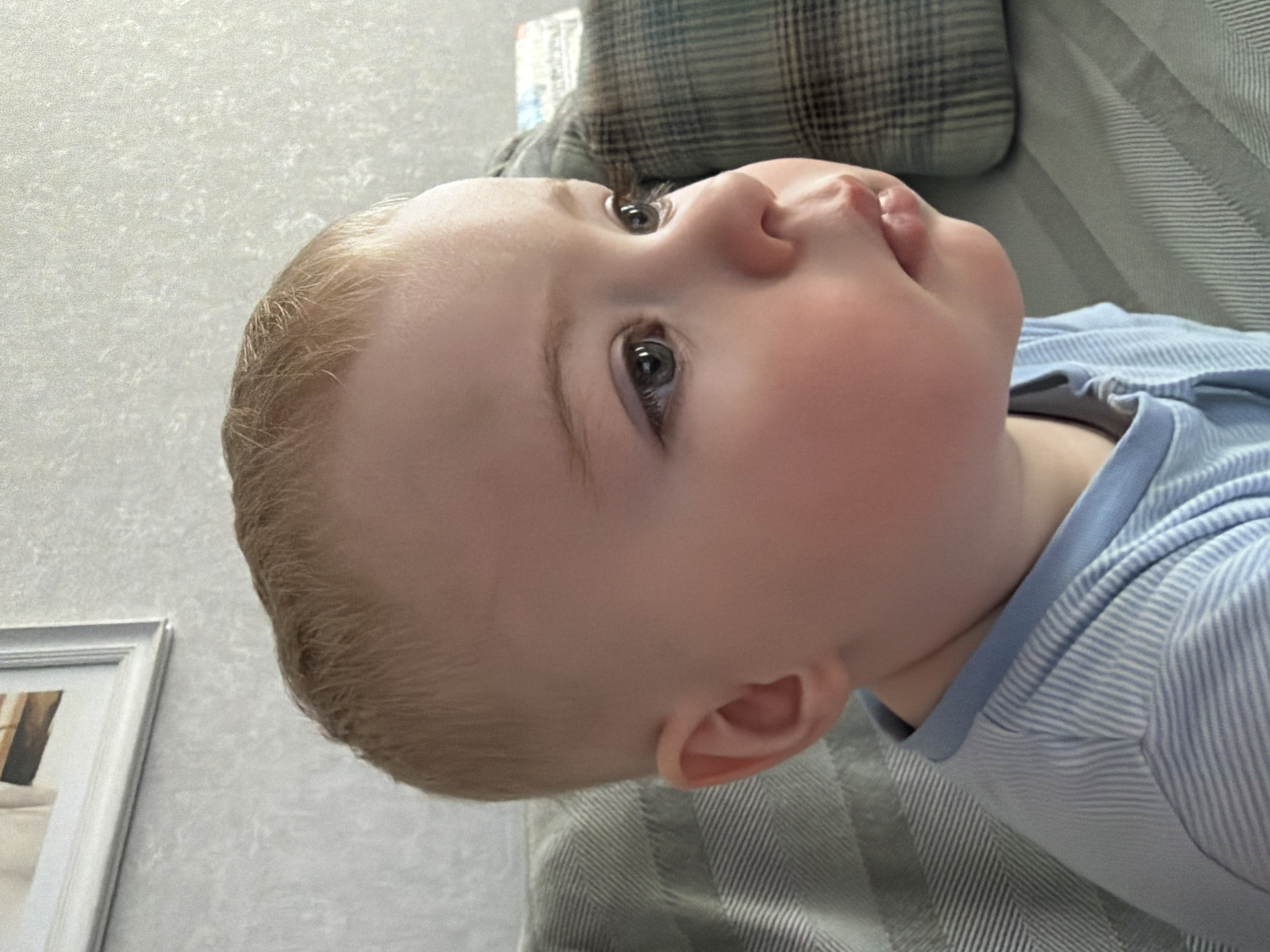 A close-up, medium-angle shot captures a young child, possibly an infant or toddler, lying on what appears to be a patterned couch or bed. The child has fair skin, light blonde hair, and grey eyes. They are wearing a light blue and white striped garment. Their head is turned upwards and to the side, with their face in profile, allowing a clear view of their cheek, nose, and slightly parted lips. The background includes a wall with a textured surface and a framed picture visible in the lower left corner. The scene appears to be indoors, possibly a residence. There is no information to confirm this photograph was taken in Votkinsk, Russia.