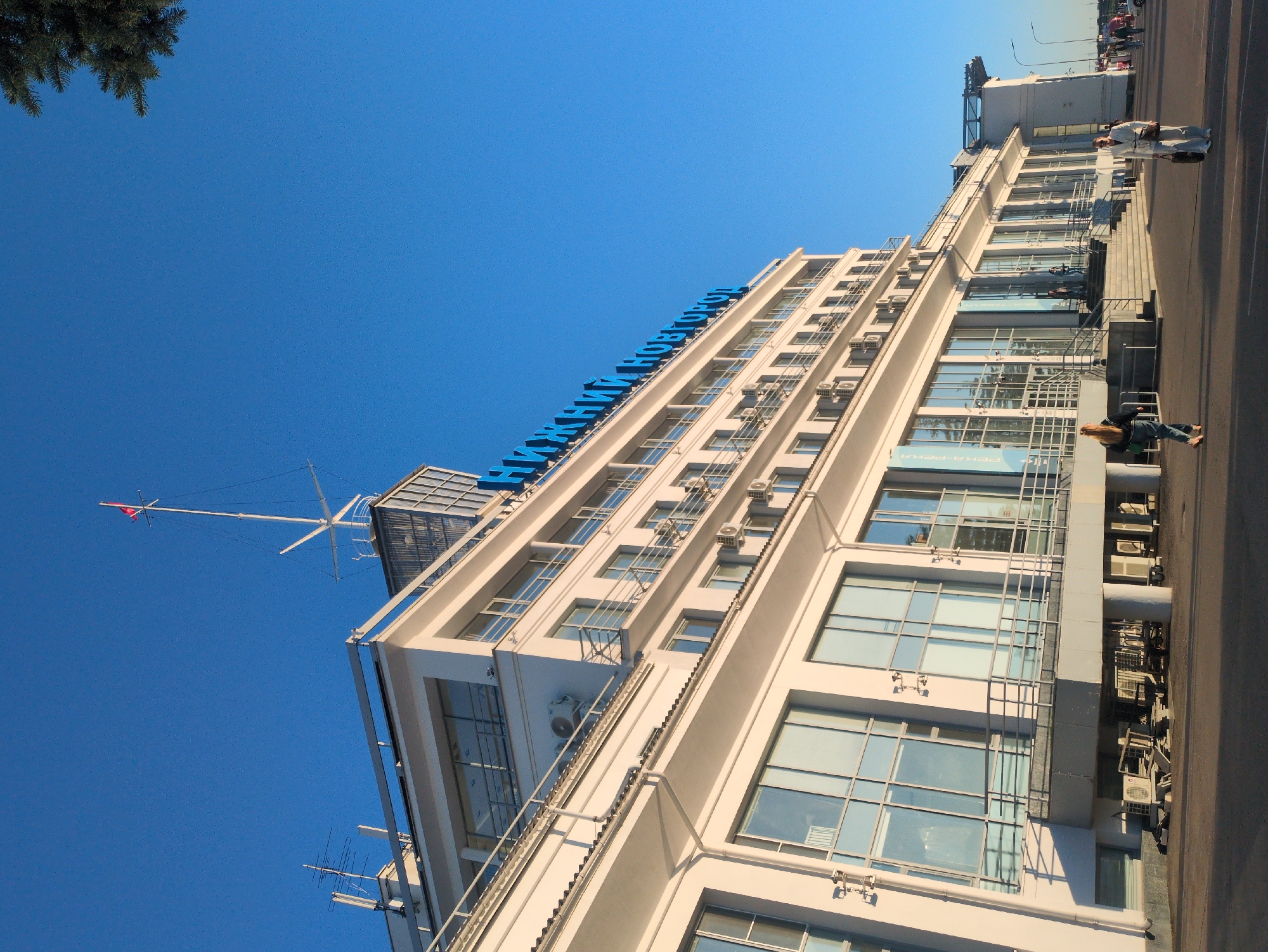 A bright, sunny day is depicted in Nizhny Novgorod, Russia, with a clear blue sky dominating the scene. The camera is tilted upwards, focusing on a modern, multi-story white building. The building has numerous large windows, many of which reflect the sky and surrounding structures. Air conditioning units are visible mounted on the exterior walls of the building.
A large, blue sign with Cyrillic lettering is mounted along the upper edge of the building, spelling out "НИЖНИЙ НОВГОРОД" (NIZHNY NOVGOROD). To the left of the building, a flagpole with a red flag is visible against the blue sky. The image also shows a portion of a tree with dark green needles in the upper left corner, suggesting there might be a park or green space nearby.
On the sidewalk in front of the building, several people are present. Two individuals, one in a white outfit and the other in dark clothing, are standing near the building entrance, appearing to converse. Further down the sidewalk, another person in dark clothing is walking away from the camera. The overall impression is of a typical urban environment on a pleasant day. The time of day appears to be late afternoon, indicated by the long shadows cast on the ground.