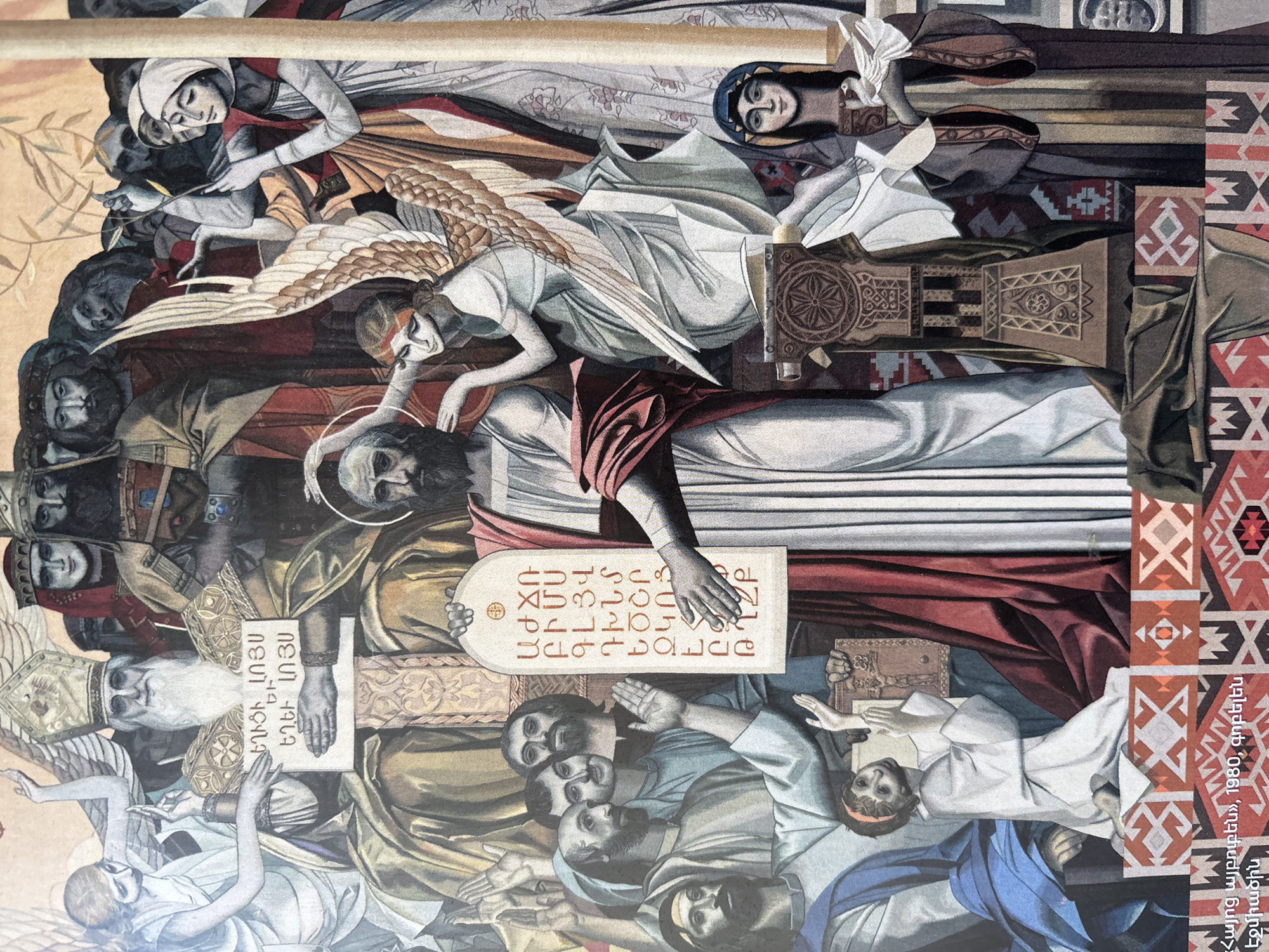 This is a depiction of the creation of the Armenian alphabet. It is a tapestry, made in 1980, called "Hayots Aybuben" (Armenian Alphabet). The location is Etchmiadzin (also known as Vagharshapat) in Armenia.
The image shows several people, most of whom appear to be male figures. The central figure has a halo above their head and seems to be lying horizontally, supported by an angel. Some figures hold tablets inscribed with Armenian letters. There are also figures wearing miters, indicating bishops or clergy.
The image contains objects such as a wooden panel with an ornate carving and another section filled with traditional Armenian textile patterns. The action taking place shows figures creating the Armenian alphabet.