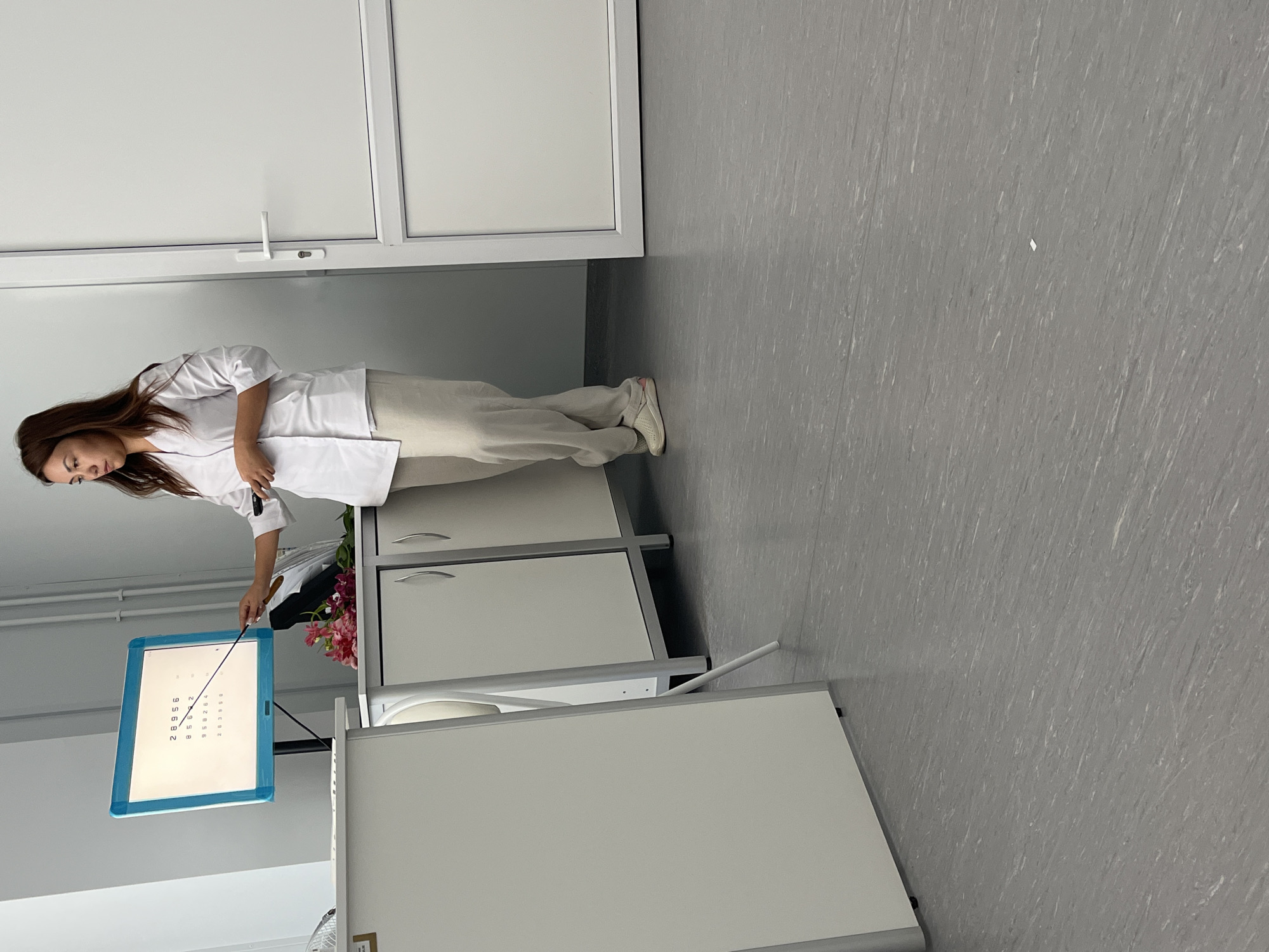 A woman in a white lab coat and beige pants is standing in front of a computer monitor displaying rows of numbers, holding a pointer in her right hand. She appears to be demonstrating something on the screen, as her gaze is directed towards it, and the pointer is extended towards the display. To her left, a bouquet of pink and red flowers rests on a white cabinet. The room has light gray flooring and white walls, with a closed white door to the right. The scene is indoors, likely a medical or educational setting, possibly an optometry clinic, given the eye chart display on the monitor. There are no other individuals visible in the frame. The lighting is bright and even, suggesting an indoor environment with artificial illumination. The overall impression is a professional setting where an examination or presentation is taking place.
