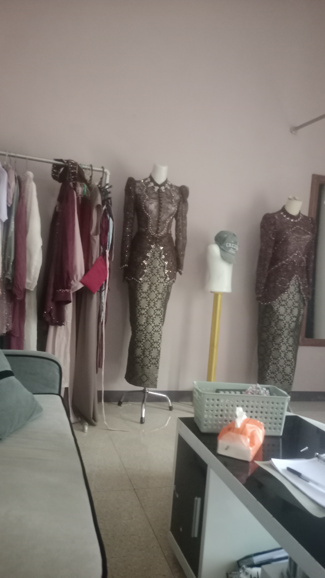 The image depicts an indoor scene, likely a room within a dress shop or a tailor's studio in Pakemitan, Indonesia. In the foreground, a portion of a light-colored sofa with a dark trim is visible on the left. A low, modern cabinet with a dark top and a white and black base occupies the right side of the image. On top of the cabinet, there is a light green basket containing some items, and an orange plastic bag with white tissues inside. A few papers and a pen are also on the cabinet's surface.
The background reveals a clothing rack with several garments hanging on it, predominantly in muted colors like white, pink, and burgundy. To the right of the clothing rack are two mannequins dressed in traditional Indonesian attire. The mannequin on the left wears a detailed brown and gold patterned long dress with a high neckline and puffed sleeves, adorned with sequins and beading. The mannequin on the right is similarly dressed in a dark maroon and gold patterned outfit. Between the two mannequins, a third, smaller white mannequin displays a green baseball cap. The walls are painted a neutral, light pinkish hue. The floor appears to be tiled with a glossy finish. The overall impression is that of a display or storage area for formal wear.