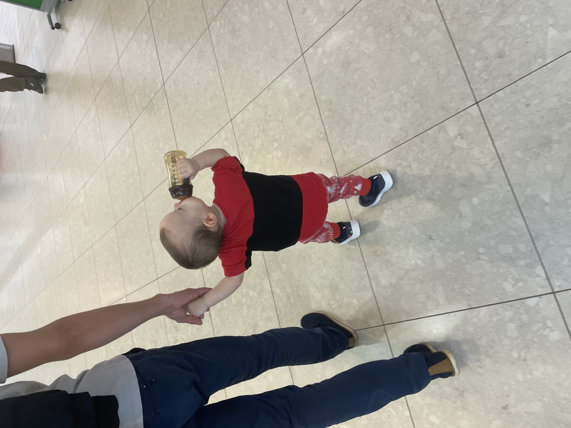 A toddler, wearing a red and black shirt, patterned red and white pants, and blue and white sneakers, lies on a light-colored tiled floor, drinking from a bottle. An adult holds the toddler's hand. The adult wears blue pants and brown shoes. The setting appears to be an interior public space with tiled flooring. It is not possible to identify the city of Perm, Russia, from the visual content.