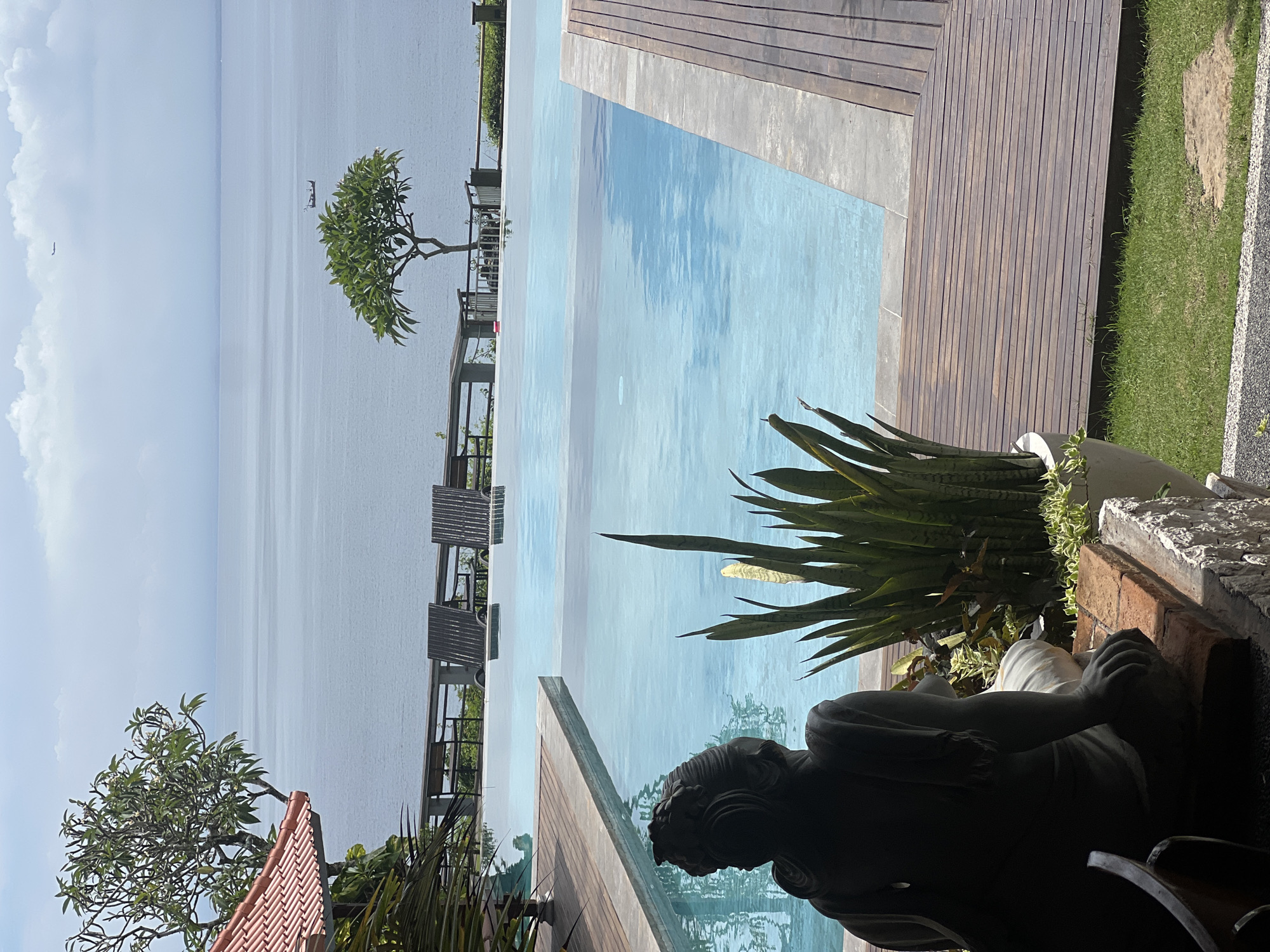 The image shows an outdoor scene at a resort in Kuta Selatan, Indonesia. An infinity pool extends toward the ocean. Wooden decking and grass borders the pool on one side. A statue of a reclining person and several potted plants are positioned beside the pool. Several chairs are placed at the edge of the ocean with a tree providing shade. In the distance, a seemingly endless view of the ocean fades into a cloudy sky.