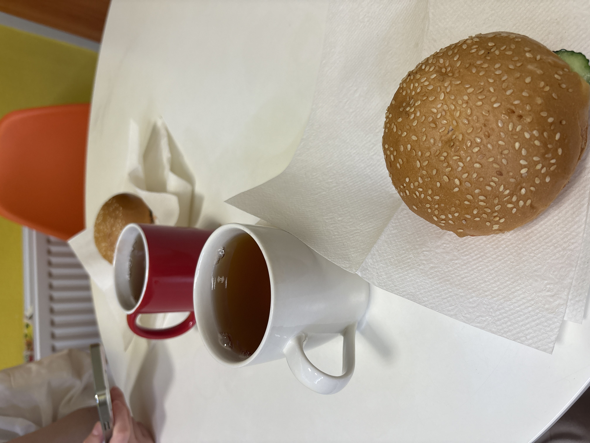 The image shows a white table in Sinyavino, Russia, on which are two mugs filled with a brown liquid, likely tea. One mug is red and the other is white. To the right of the mugs is a hamburger bun with sesame seeds on a white paper napkin. To the left of the mugs is a smaller round bun on a napkin. A person’s hand holding a smartphone is visible in the bottom left corner of the frame. An orange chair and a yellow wall are visible in the background, to the left.