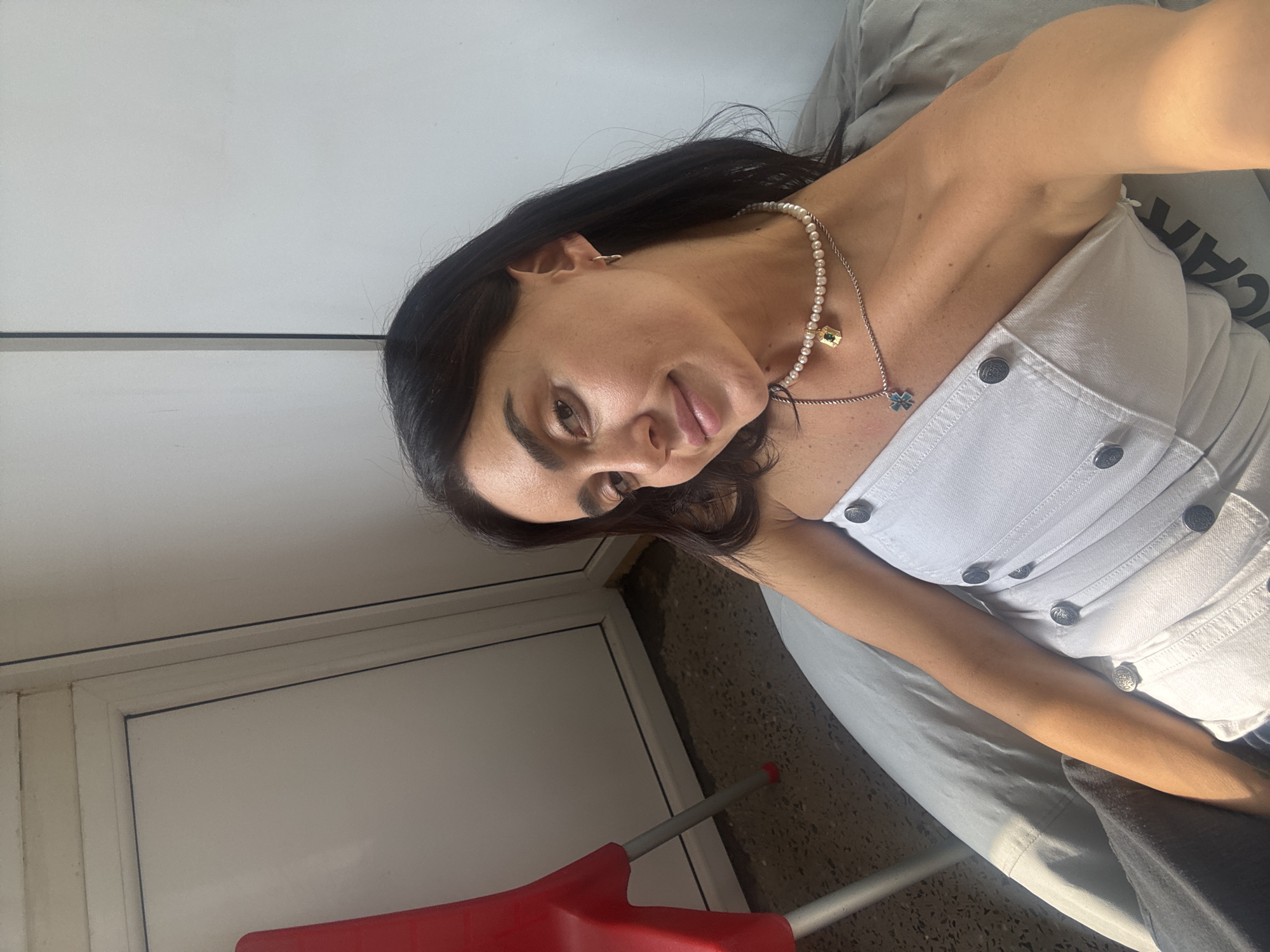 A young woman with dark hair and brown eyes is pictured in a selfie. She is wearing a white strapless top and a pearl necklace. She is sitting in what appears to be a beanbag chair. Behind her, to the left, is a red chair. The background is a plain white wall with a doorframe. The lighting suggests it is daytime. The location is identified as Tbilisi, Georgia.