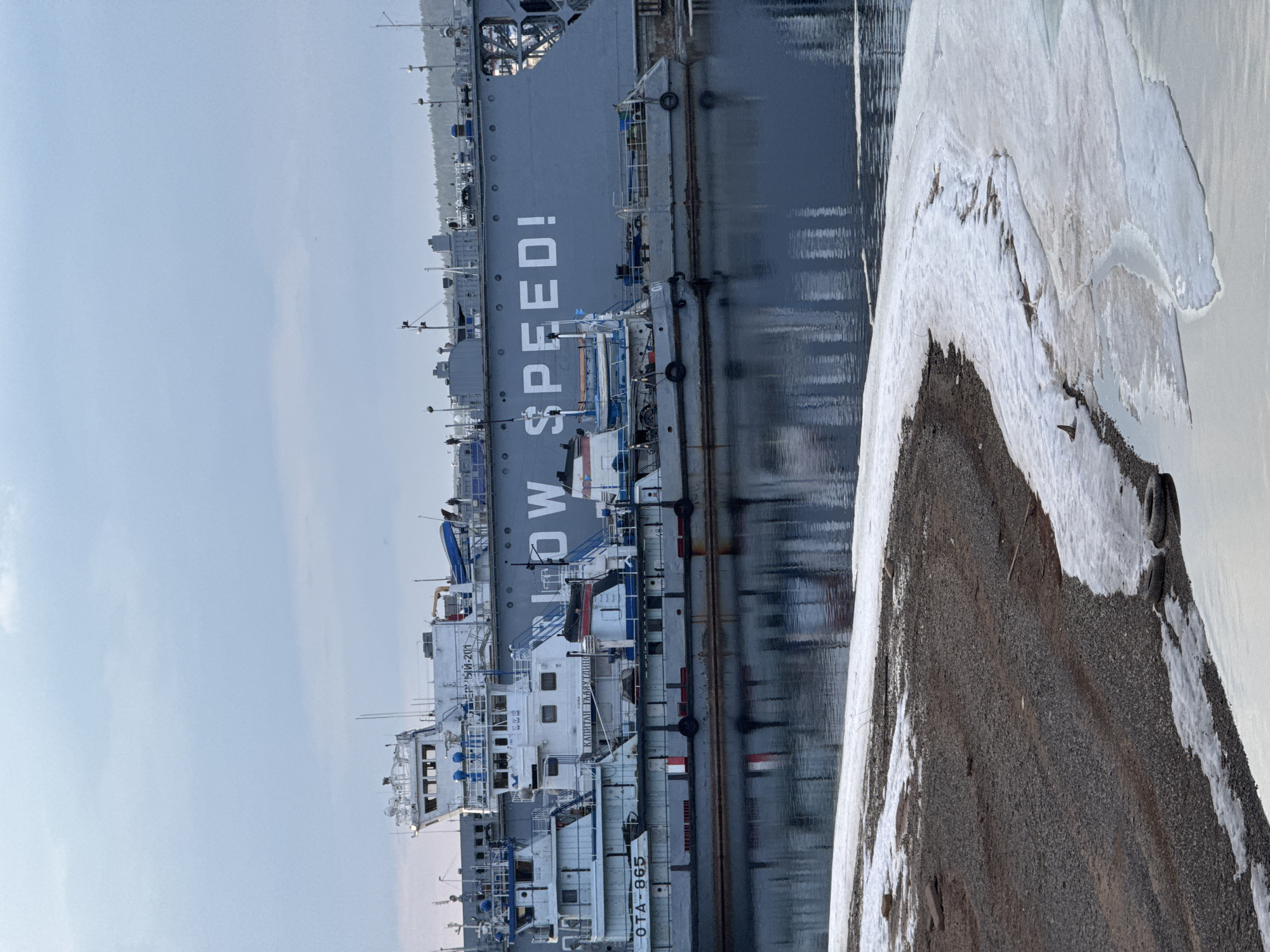 The image depicts a waterside scene featuring multiple vessels docked in what appears to be a port or industrial waterway. On the left, a large, dark grey ship is prominently displayed, with the words "LOW SPEED!" painted in large white letters on its hull. Its reflection is visible in the calm, dark water.
Alongside the large grey vessel, or docked in front of it, are several smaller white boats, likely tugboats or service vessels. Visible text on one of these smaller boats includes "OTA-865" near its waterline. Another white boat shows the Cyrillic text "КАПИТАН ТАЙМУР" and "ГЕРМЕР 1510-201" on its superstructure.
The right side of the image features a shore composed of dark gravel or dirt, heavily interspersed with patches of white snow and ice, indicating cold weather conditions. Some discarded tires are partially visible on the icy shore. The water near the shore also contains slushy ice formations. The sky above is a clear, pale blue, suggesting daylight and fair weather despite the cold. There are no people visible. The scene captures a moment of apparent inactivity in a working port during a cold season.