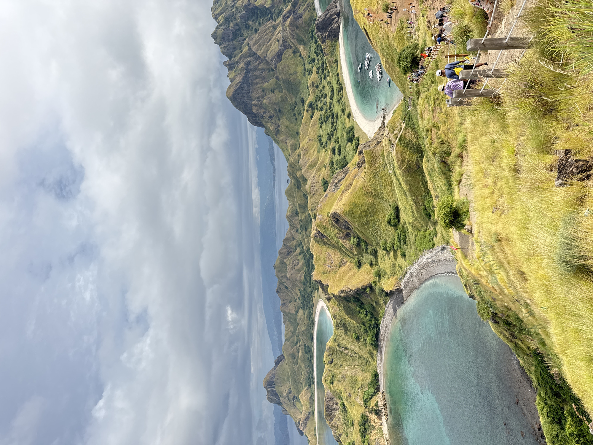 This media file captures a dramatic panoramic landscape, viewed from a high vantage point overlooking a series of rugged, undulating hills meeting the sea. The image is oriented such that the sky is on the left, and the land and sea extend to the right.
The scene features distinctive, grass-covered hills, predominantly green with patches of golden-brown, suggesting a blend of lush vegetation and drier areas. Several pristine coves with crescent-shaped white sand beaches are visible, their clear, turquoise waters dotted with multiple small boats, likely tourist vessels, anchored near the shore. One prominent cove in the middle right of the image showcases these anchored boats and beachgoers. Another, more distant cove further down the coast also appears with calm, clear waters.
On the highest ridge, which dominates the foreground on the upper right side of the image, a group of people are gathered. They are seen along a clearly marked path, some standing, some sitting, observing the expansive vista. A wooden railing runs along this elevated path, indicating a designated viewpoint or hiking trail. The people appear to be tourists or hikers, enjoying the scenery.
The sky is largely overcast with soft grey and white clouds, suggesting a cloudy but bright day. In the distance, the calm, blue waters of the ocean extend to the horizon, meeting a hazy, distant landmass or mountain range. The overall impression is one of awe-inspiring natural beauty, highlighting a majestic coastline and marine environment, likely a popular destination for sightseeing and outdoor activities. No visible text is present in the image.