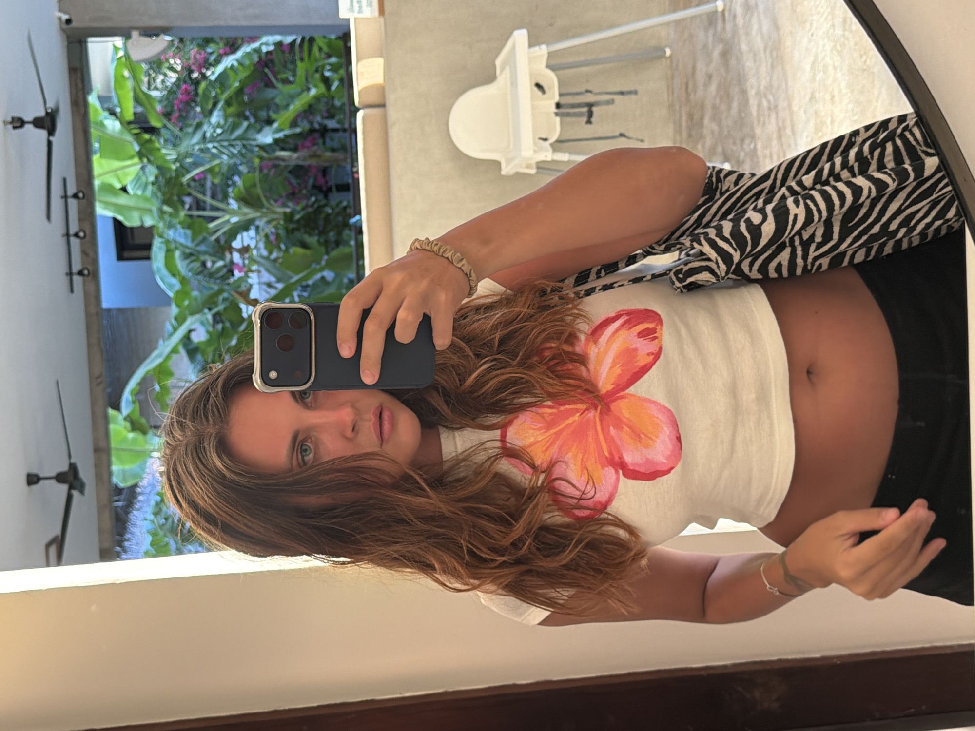 A mirror selfie captures a young woman holding a dark-colored smartphone in her right hand, positioned centrally within the frame. The image is oriented vertically but has been rotated 90 degrees clockwise.
The woman has long, wavy brown hair and blue-green eyes, looking directly at the camera with a neutral expression. She is wearing a white short-sleeved crop top featuring a large, colorful floral design, likely a plumeria or hibiscus, in shades of orange and pink. Her midriff is visible, and she is wearing dark-colored bottoms, possibly black shorts or pants. She has a beige scrunchie on her right wrist and a thin, light-colored bracelet on her left wrist.
The reflection reveals an indoor setting with light-colored walls or concrete surfaces. In the upper right background, a white high chair with metal legs is visible. On the left, two dark-colored ceiling fans are mounted on a white ceiling, along with some dark wall fixtures below them. Beyond an opening, a vibrant tropical outdoor scene is reflected, characterized by abundant lush green foliage, including large leaves and clusters of pink or purple flowering plants. The bright natural light suggests it is daytime.
The overall scene indicates a warm, possibly resort-like environment in General Luna, Philippines. There is no discernible text visible in the image.