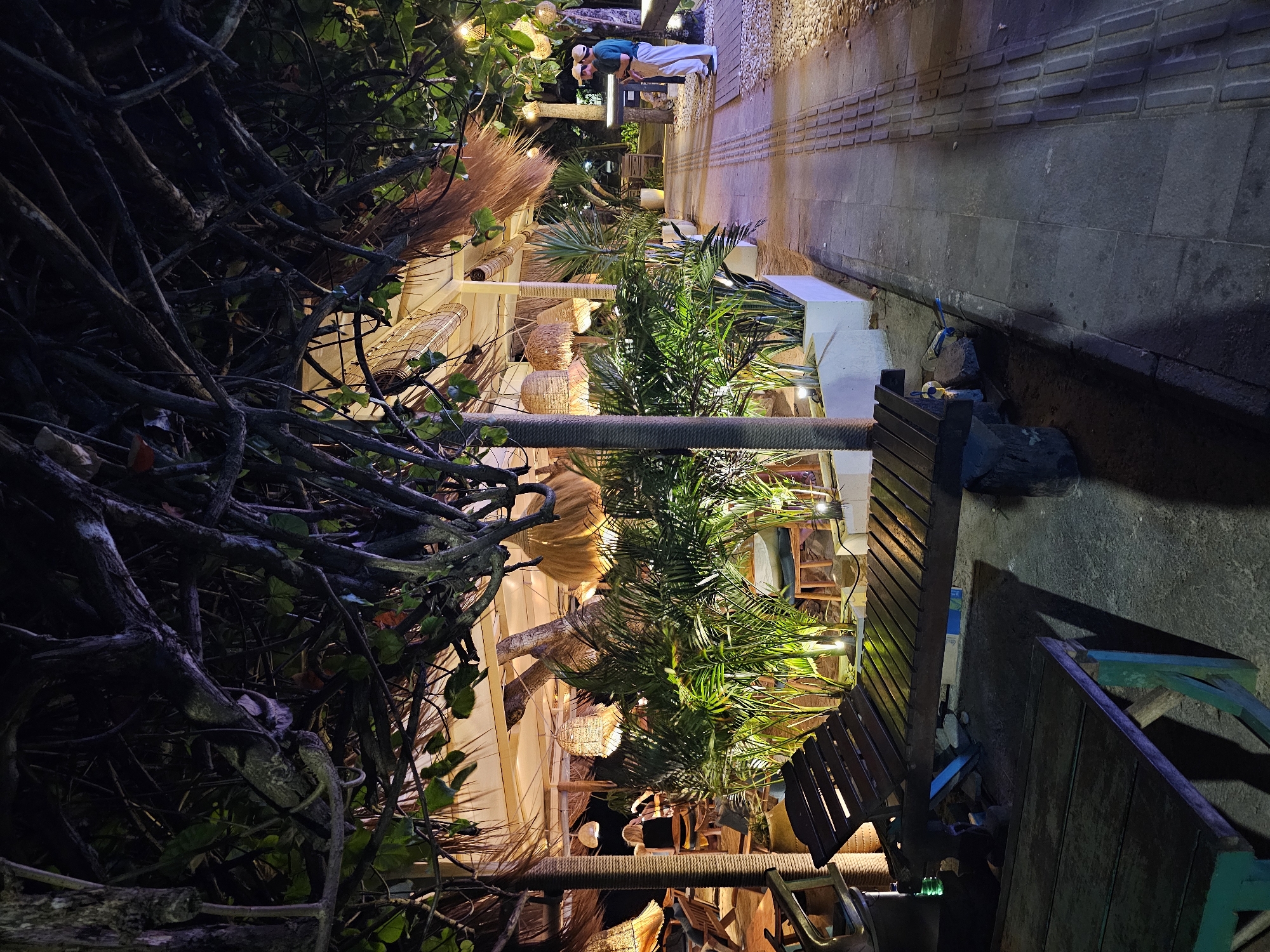 The image depicts a nighttime scene at a beachfront restaurant in Denpasar, Indonesia. The foreground features a wooden lounge chair and a portion of a wooden table, casting long shadows on the concrete and gravel pathway. Lush green potted plants with artificial lighting illuminate the area. In the mid-ground, a thatched roof structure is visible, likely part of the restaurant's seating area, with several woven light fixtures hanging from the ceiling. Two individuals, dressed in casual attire, are standing near a dark-colored structure in the right mid-ground, facing each other. The background shows a walkway lined with small stones and brick pavers, extending towards more of the restaurant's seating and vegetation. The overall ambiance suggests a tropical, outdoor dining environment illuminated by ambient lighting.