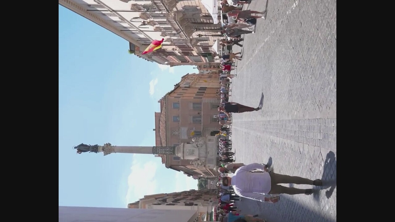 The image depicts a cobblestone street lined with buildings, a monument with a statue atop it, and a Spanish flag flying from a building. Several people are walking in various directions, some appearing to be tourists. A man in a white shirt and brown pants is walking away from the camera, carrying a drink. Another man in a dark shirt and pants is walking towards the camera, talking on his phone. In the background, more people are visible, some near the monument and others further down the street. The sky is clear and blue. The scene appears to be a public square or pedestrian walkway in an urban setting, likely a tourist area given the presence of a monument and people with a leisurely demeanor. The city of Corchiano, Italy, is mentioned as the location, suggesting this is a prominent landmark or street within that town.