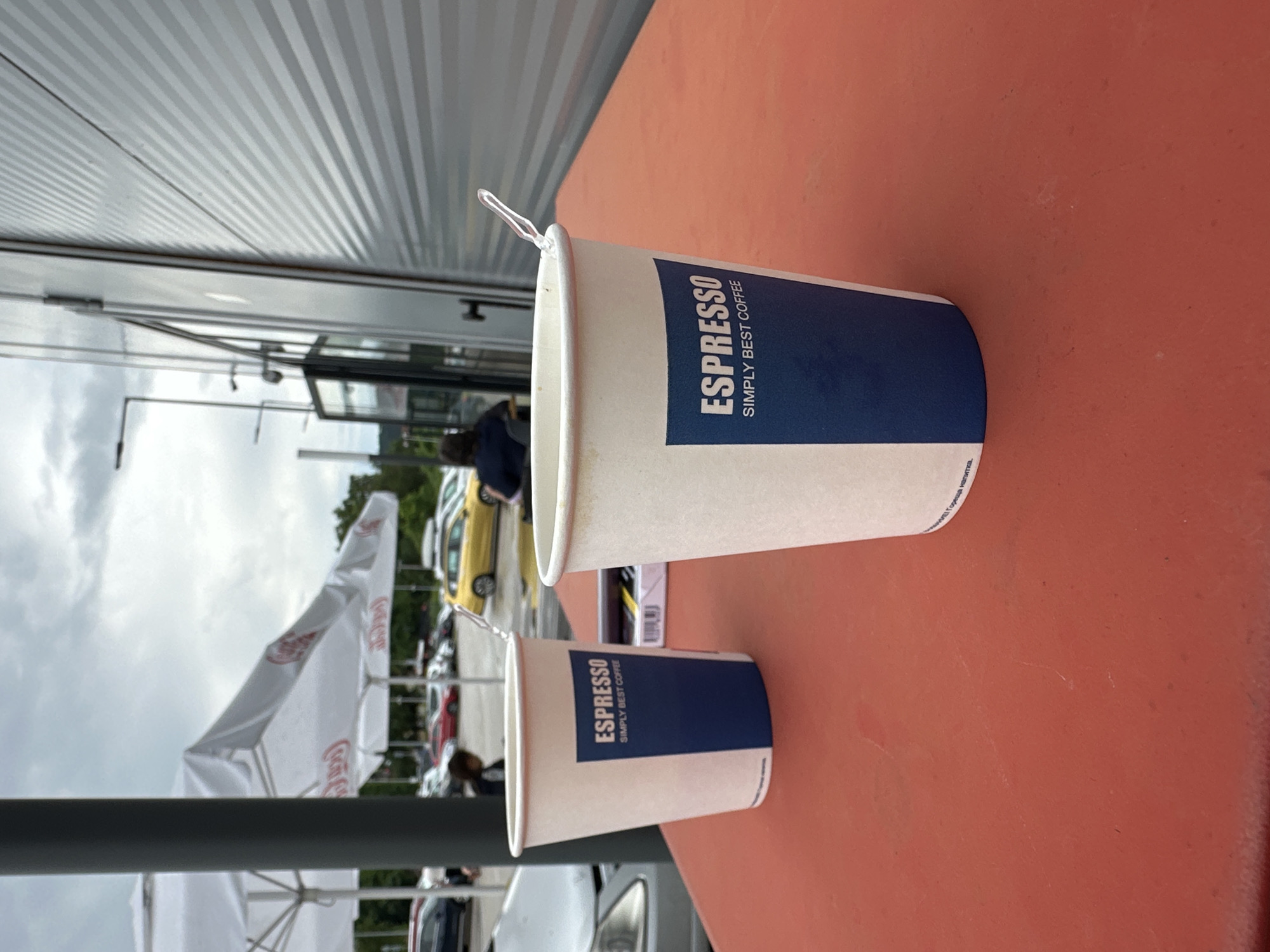 This outdoor image, captured in Lovech, Bulgaria, depicts a casual coffee stop under an overcast sky. In the foreground, a prominent reddish-orange table stretches across the lower half of the frame. On the table are two white paper coffee cups, differing in size. Both cups feature a distinctive blue label displaying "ESPRESSO" in large white capital letters, with "SIMPLY BEST COFFEE" written underneath in smaller white text. A clear, slender plastic stirrer or spoon protrudes from the rim of the larger coffee cup.
The background reveals a commercial setting. To the left, a building with a horizontally corrugated metallic exterior is visible. Further back, a large white umbrella, likely branded with red "Coca-Cola" text, provides shade. Several cars, including a noticeable yellow vehicle, are parked in the distance. The partial figures of people can be seen; one person wearing a dark blue shirt is visible near the cars and building, with other individuals appearing as blurry forms further back. The scene suggests a daytime setting, possibly a break for coffee at a roadside establishment or cafe, characterized by the cloudy weather.