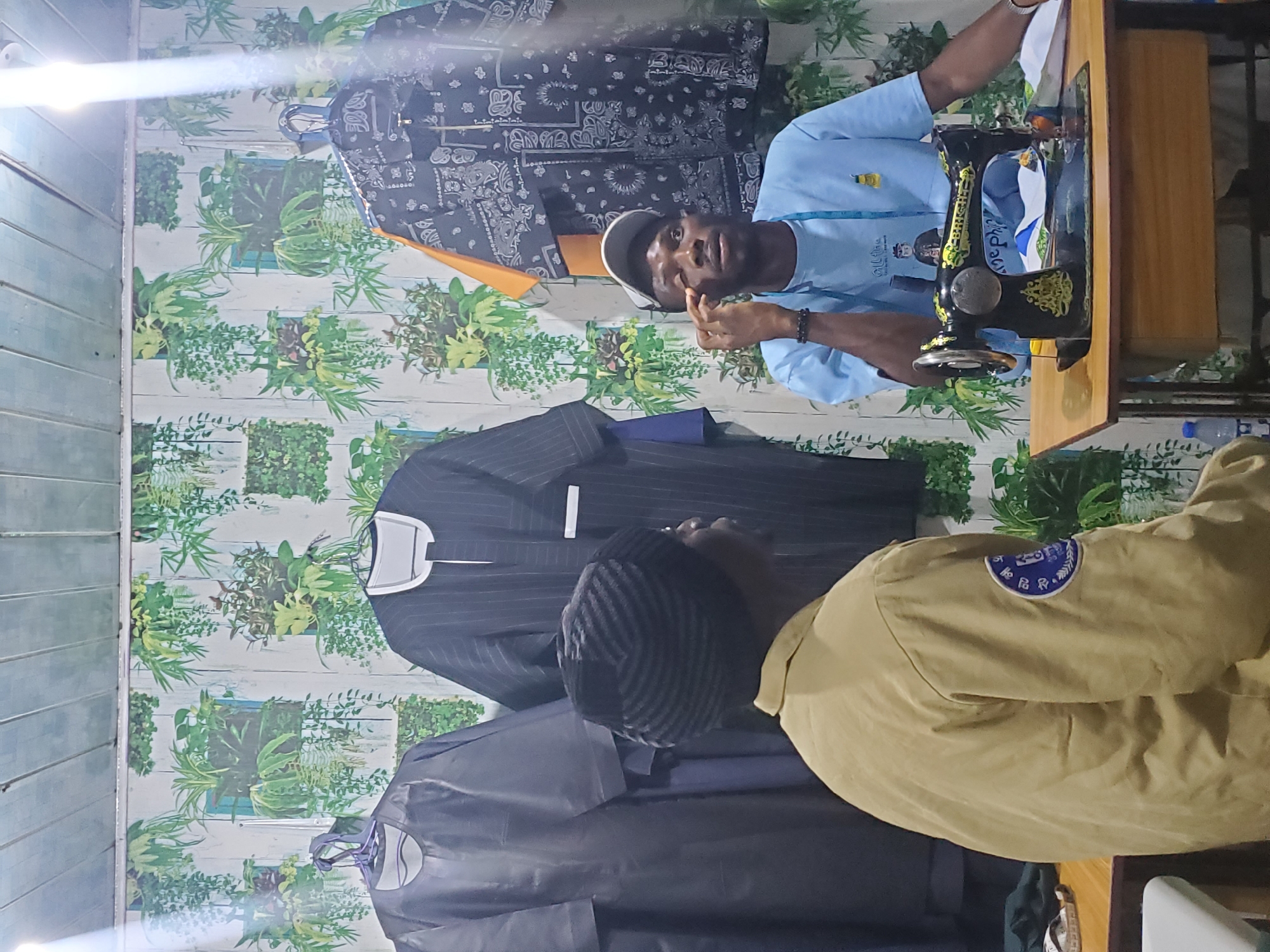 The image shows a tailor working on a sewing machine in a workshop. The tailor, a young man wearing a light blue shirt and a white cap, is looking at the camera and appears to be speaking. He is positioned behind a vintage black and gold sewing machine. The workshop has a patterned wallpaper featuring a botanical design with green foliage. Various garments, including a pinstripe suit and a dark grey garment, are hanging in the background. Another person, partially visible in the foreground, is wearing a tan uniform with a blue and white emblem on the sleeve. The emblem contains text in a circular design, but it is not clearly legible. The lighting suggests it is daytime, and the overall impression is of a busy tailoring establishment. The location is identified as Jos, Nigeria.