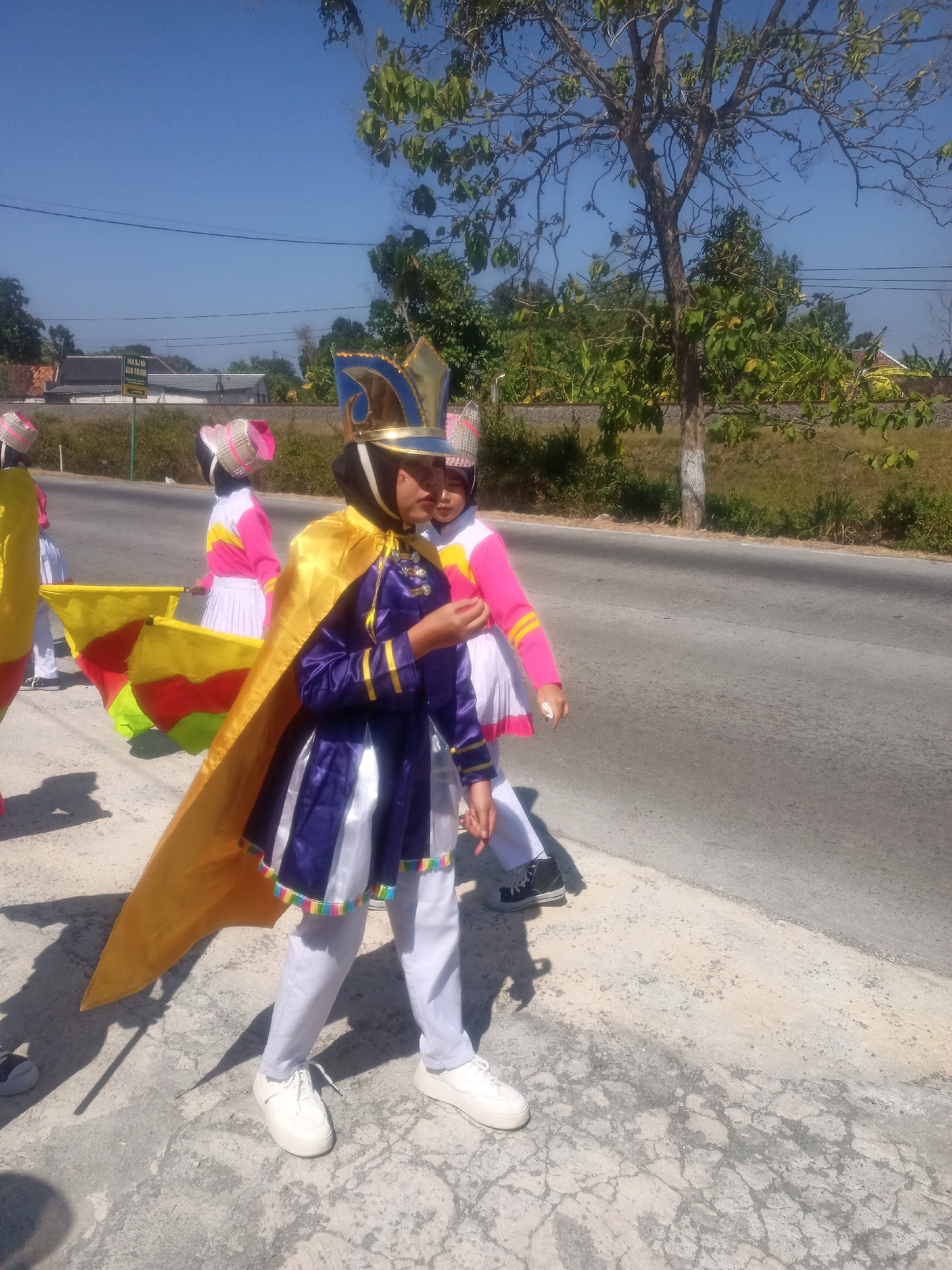 This image depicts a group of children dressed in colorful marching band uniforms and elaborate hats, seemingly participating in a parade or performance on a sunny day. The children are positioned along a paved road, with a grassy area and trees to the right. In the background, there are some buildings and a sign that reads "MASJID ASA-SIHUL". This suggests the location is in Sumberlawang, Indonesia, where a mosque named ASA-SIHUL is present.
The central figure in the foreground wears a purple and white uniform with a golden cape and a decorative blue and gold crown-like hat. They are holding a flag that displays red, yellow, and green colors. To their left, another child in a yellow uniform is partially visible. To the right of the central figure, another child in a pink and white uniform and a similar hat is also visible.
The overall scene is bright and airy, with clear blue skies and abundant sunlight casting shadows on the ground. The road appears to be an asphalt surface, and the surrounding vegetation suggests a somewhat rural or suburban environment within the city. The children's attire and the flag indicate a festive and organized event.