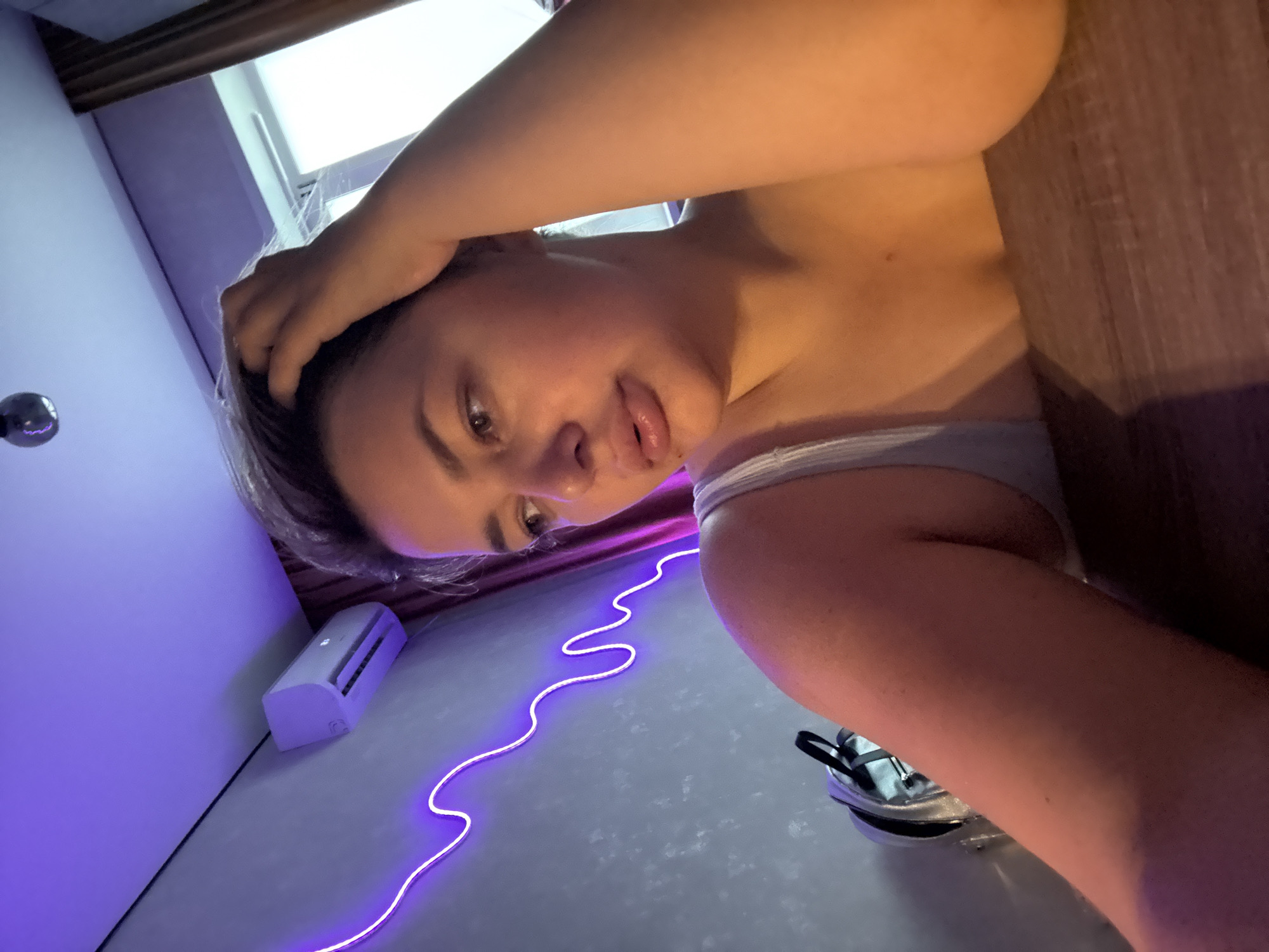 In Birobidzhan, Russia, a woman is captured in a close-up, low-angle shot, seated at a wooden table. She has short, light brown hair and is wearing a light-colored tank top. One hand is raised to her head, while her gaze is directed downwards and to the left. The background features a light grey wall with a winding neon purple light fixture. To the left, a white air conditioning unit is mounted high on the wall. A portion of a silver bag with black straps is visible on the floor near the wall. The lighting is dramatic, with the purple neon and ambient light casting shadows and highlighting the woman's features. The overall impression is a dimly lit, modern interior space.