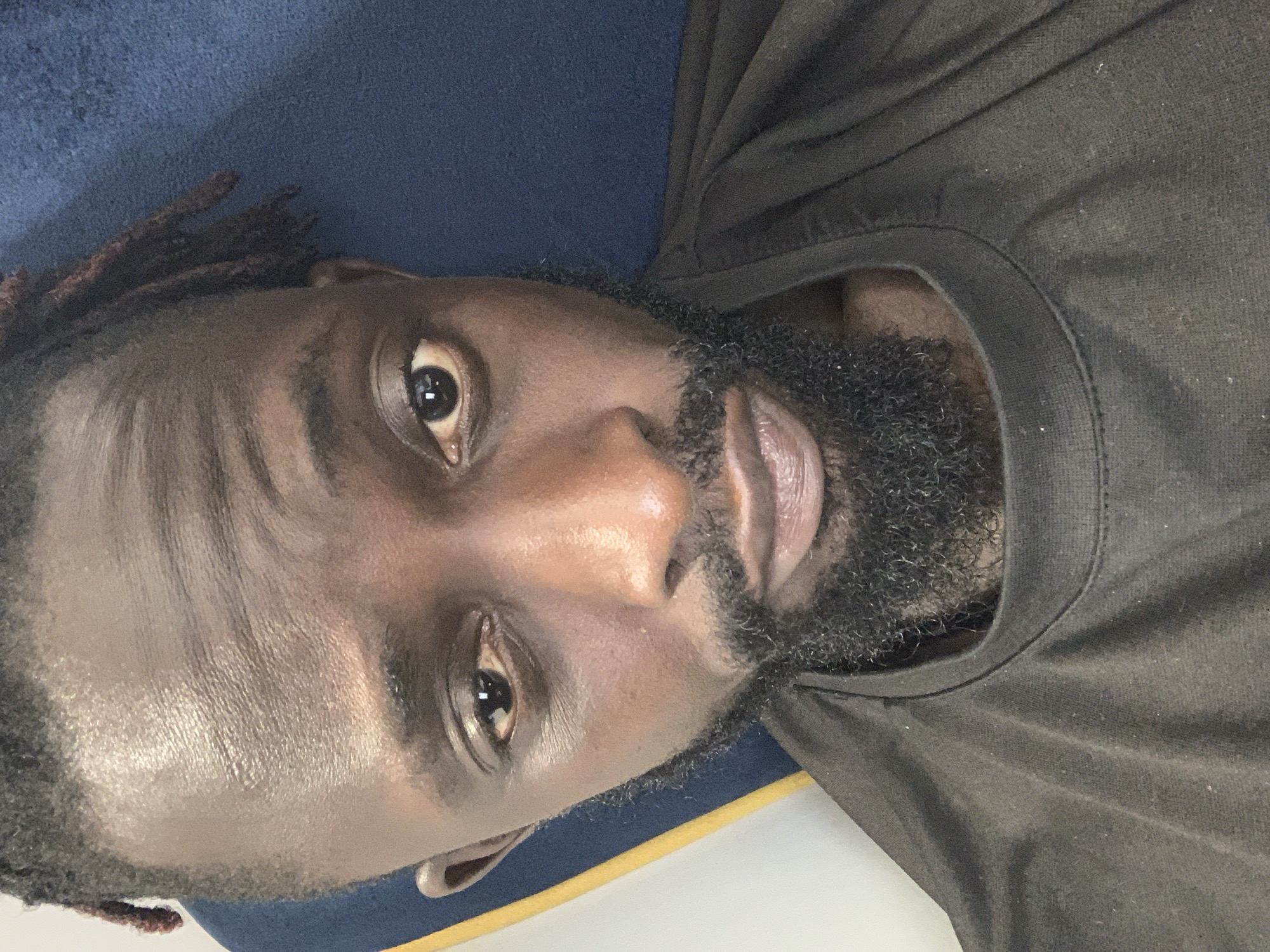A close-up, angled portrait shows a dark-skinned man with a black beard and dreadlocks. He is wearing a dark brown t-shirt. The background includes a blue surface and a white area with a yellow stripe. There is no information provided in the image about the location being in Lekki, Nigeria.
