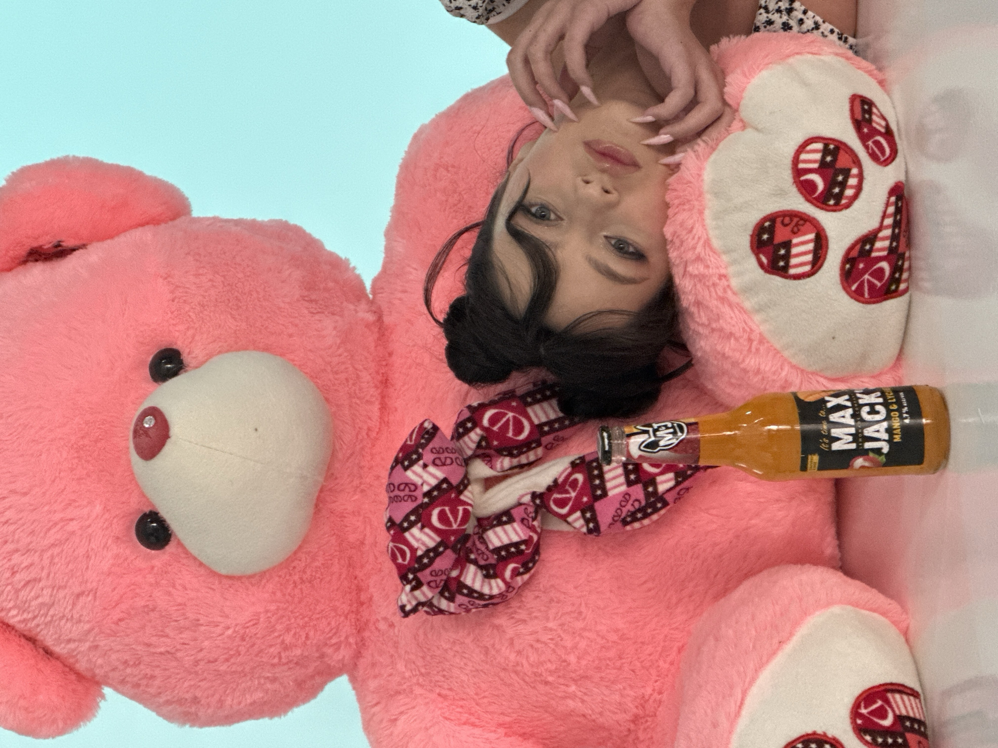 A woman with dark hair in buns is lying on a large, pink teddy bear, holding a bottle of Max Jack mango and lychee flavored alcoholic beverage. The teddy bear has a white face with black eyes and a red nose. It also has a bow with red and white patterns. The location is not discernible from the image.