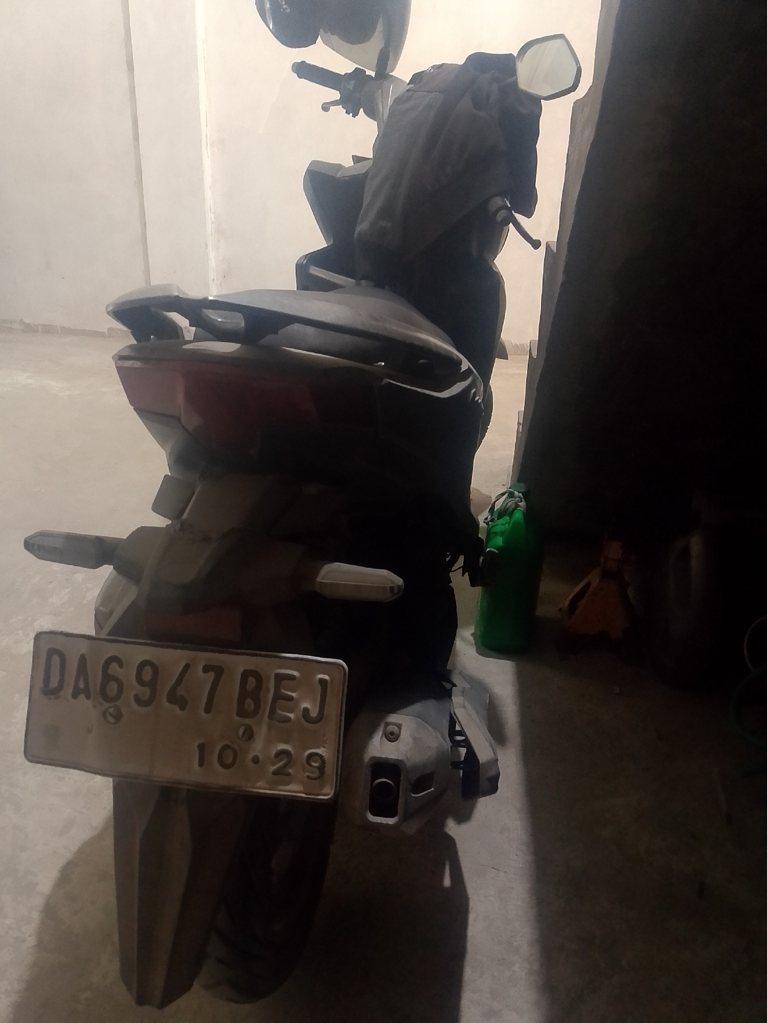 A rear-view of a dark-colored scooter is visible in an indoor, likely storage area, possibly a garage. The scooter has a license plate that reads "DA6947BEJ" with the date "10.29" below it. A dark-colored cloth is draped over the rear of the scooter, obscuring some of its details. To the right of the scooter, a green plastic jerrycan is standing upright. Further to the right, in the shadow, a small orange object with a handle is visible, possibly another container. The floor appears to be concrete. The lighting is dim, with a single light source casting shadows. The scene is devoid of any human presence or activity. The context of Banjarmasin, Indonesia, is not directly evident from the visual information in the image.