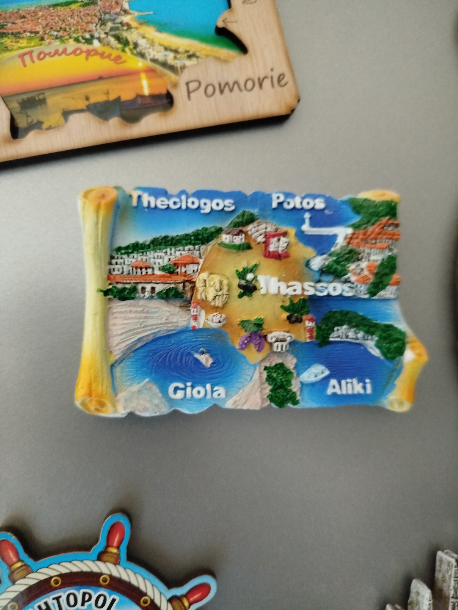 The image shows three magnets on a gray surface. At the top is a rectangular magnet, possibly made of wood, depicting the city of Pomorie, Bulgaria. The text "Pomorie" is visible on the magnet. Below that, is a map-shaped magnet representing the island of Thassos, Greece. Several locations are labeled on the magnet, including "Theologos," "Potos," "Giola," and "Aliki." At the bottom left is a partial view of a magnet with a nautical theme, featuring the word "HTOPOl" (possible typo of Sozopol, a Bulgarian city).