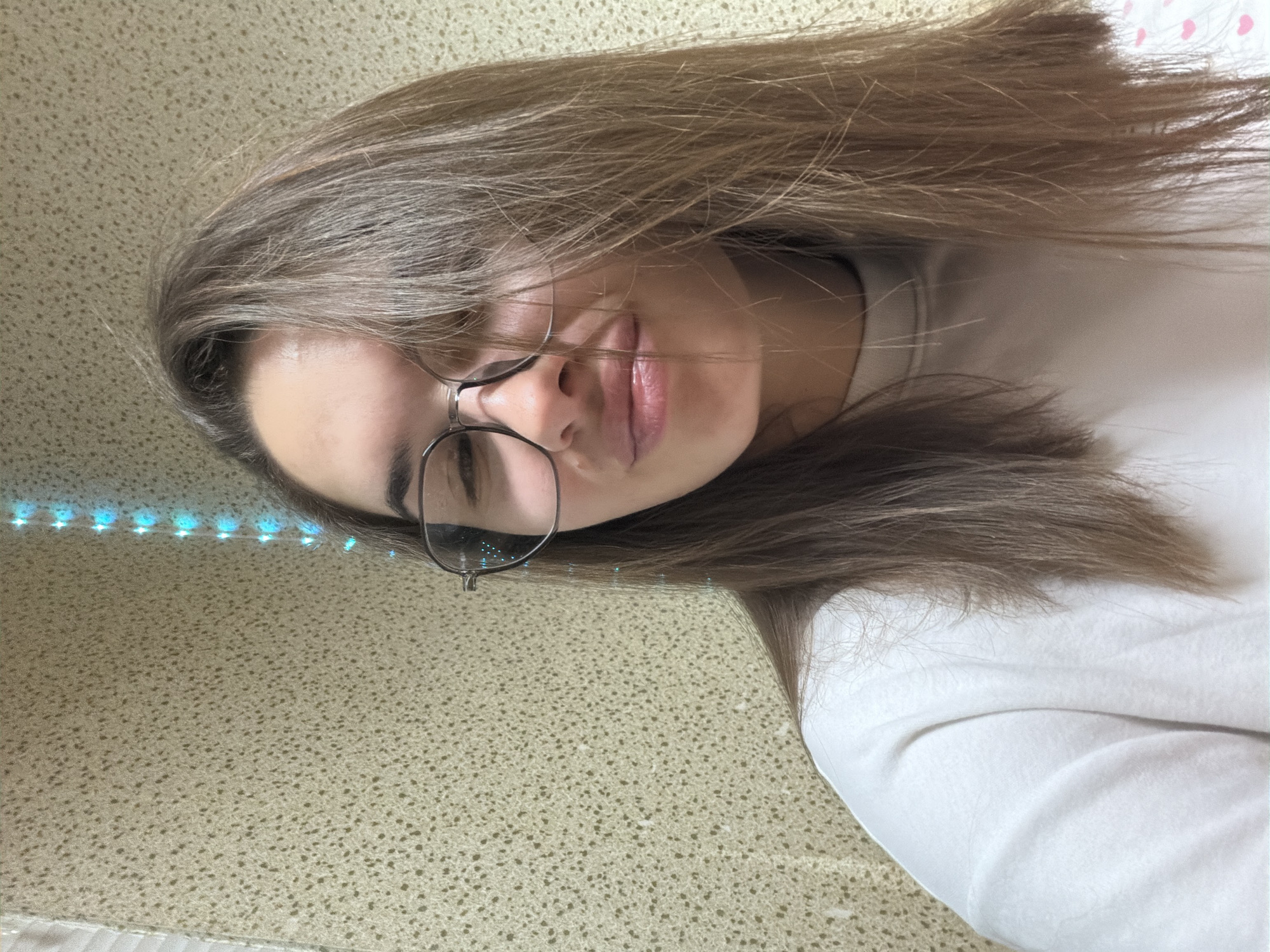 A young woman with light brown hair and glasses is pictured in a close-up shot. She wears a white top. The background is a patterned surface, likely a countertop, and a strip of blue LED lights is visible. There is no information in the image to determine if the photo was taken in Sofia, Bulgaria or any other location.