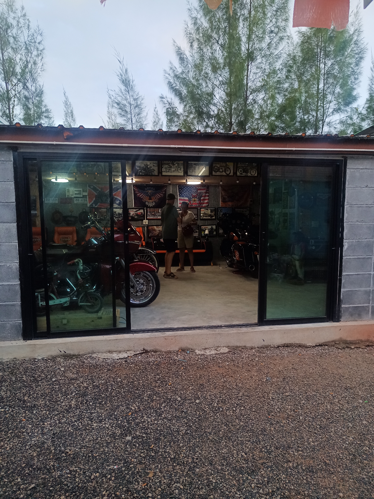 The image shows the exterior of a building in Surat Thani City Municipality, Thailand. The building is constructed of cinder blocks, with a wide opening containing sliding glass doors. Inside the building, several motorcycles are visible, along with chairs, framed pictures, and flags displayed on the back wall. Two people are standing inside the building. The ground outside the building is covered in small gravel. Trees are visible in the background. The sky is overcast.