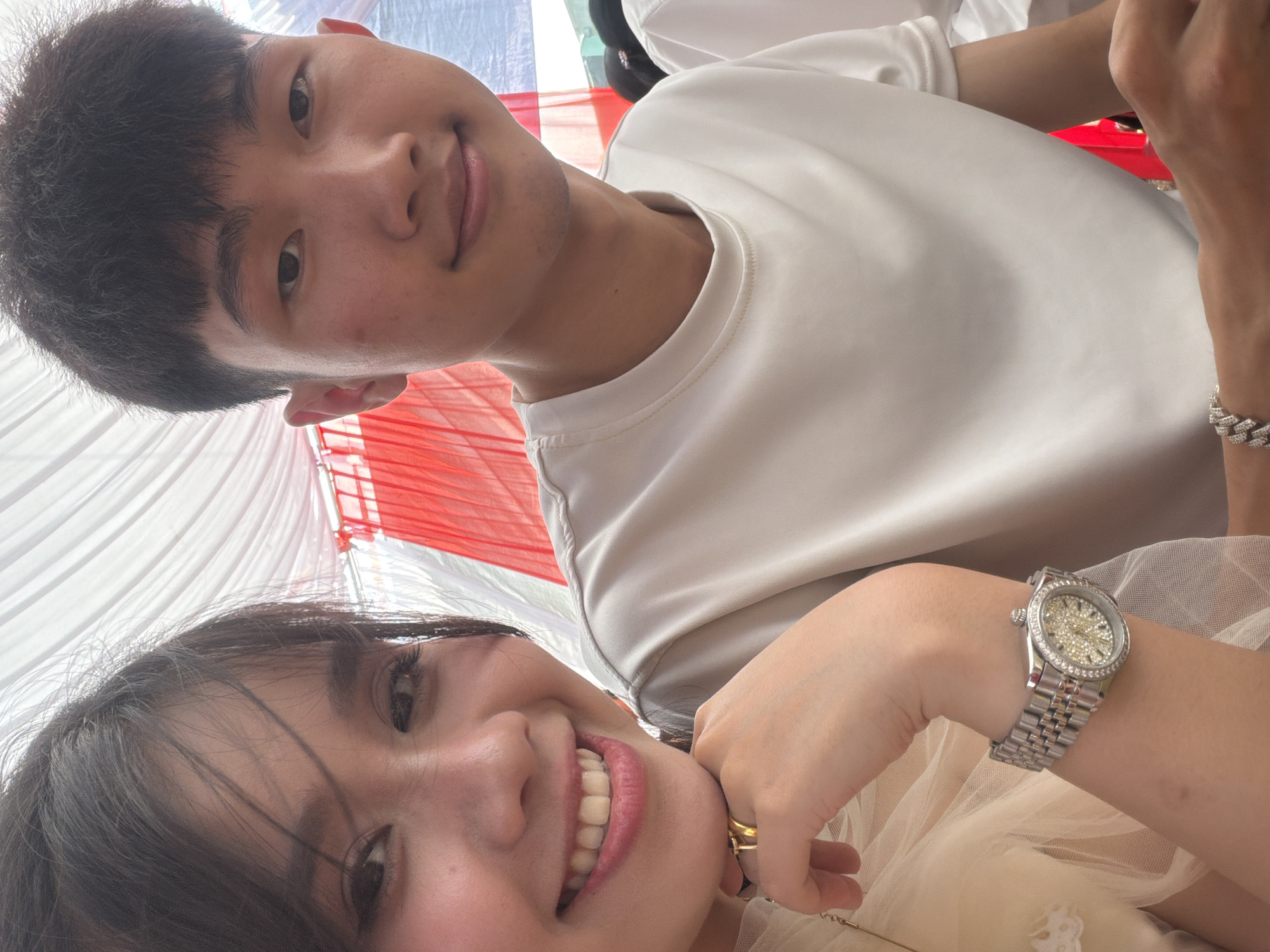 A close-up, high-angle photograph captures two young adults, a male and a female, in what appears to be a festive outdoor or semi-outdoor setting in Tay Son Commune, Vietnam. The image is a selfie, taken from below, looking upwards.
In the foreground, a female with long dark hair is smiling broadly, revealing white teeth. She is wearing a light-colored, possibly cream or beige, garment with a sheer overlay, suggesting a formal dress or top. Her left hand is positioned near her chin, adorned with multiple gold rings on one finger. On her left wrist, she wears a silver-toned wristwatch with a crystal-studded dial and bezel, and a matching metallic link bracelet.
Behind her, partially visible, is a male with short, dark hair, smiling subtly towards the camera. He is wearing a plain, light-colored t-shirt, possibly white or light grey.
The background is dominated by draped fabric in alternating red and white panels, likely part of a canopy, tent, or celebratory decoration. These colors strongly evoke the national flag of Vietnam, aligning with the given location context. The lighting suggests it is daytime. Both individuals appear happy and joyful. No visible text is present in the image.