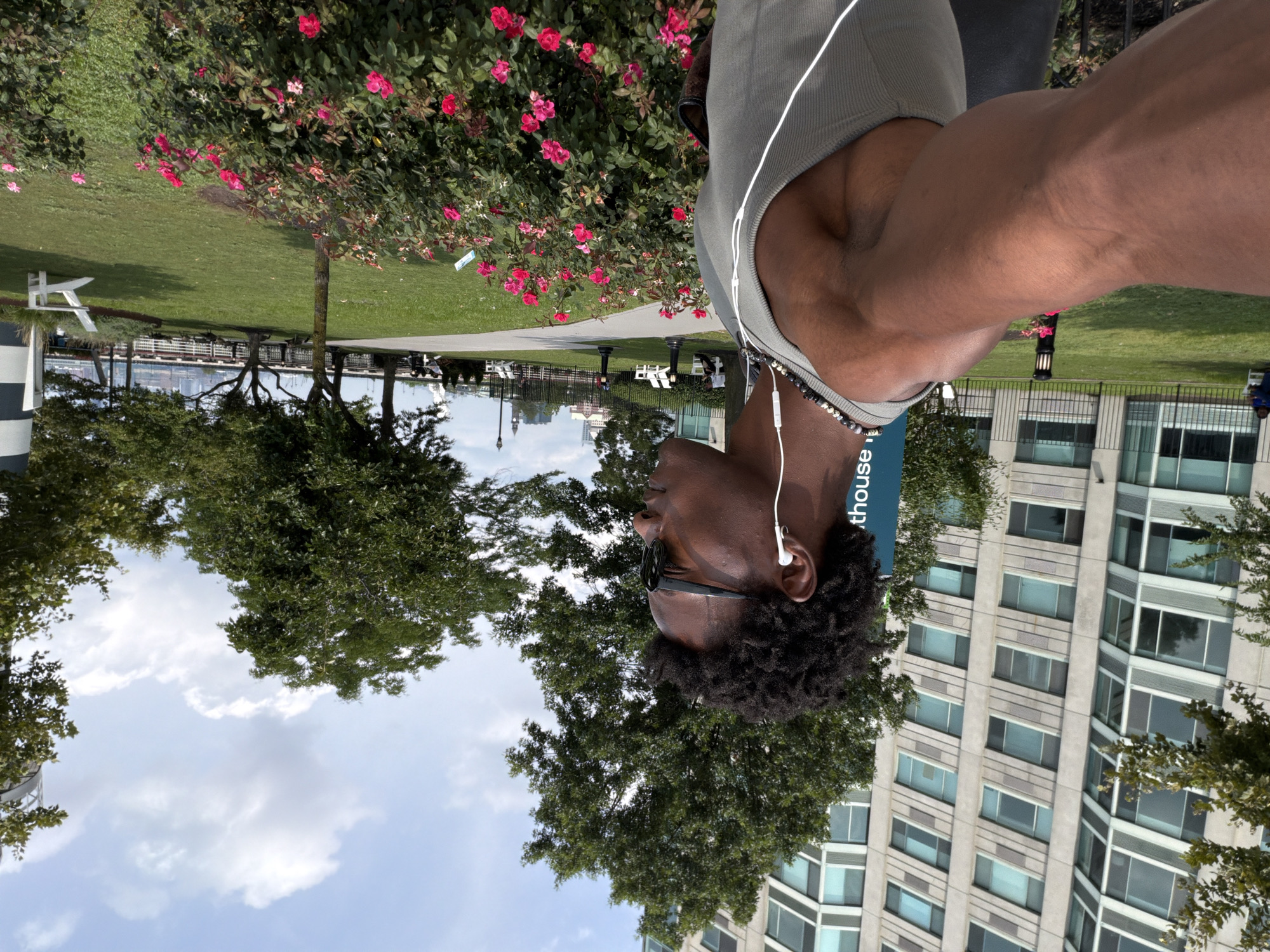 A medium shot captures a young Black man with dark, curly hair, wearing sunglasses and white earbuds, from the chest up. He is facing to the right, his left arm is extended towards the camera, and he is wearing a grey tank top and a beaded necklace. He stands in front of a bush with pink and red roses. Behind him, a large, modern apartment building with many windows dominates the background. To the right of the building, trees and a bright blue sky with white clouds are visible. The man is positioned in a park-like setting with a manicured lawn, trees, and a black fence. In the distance, several white lounge chairs and lampposts are scattered across the grass. The overall scene appears to be a sunny day in Jersey City, New Jersey, likely in a waterfront park, given the hint of buildings and a body of water in the far background.