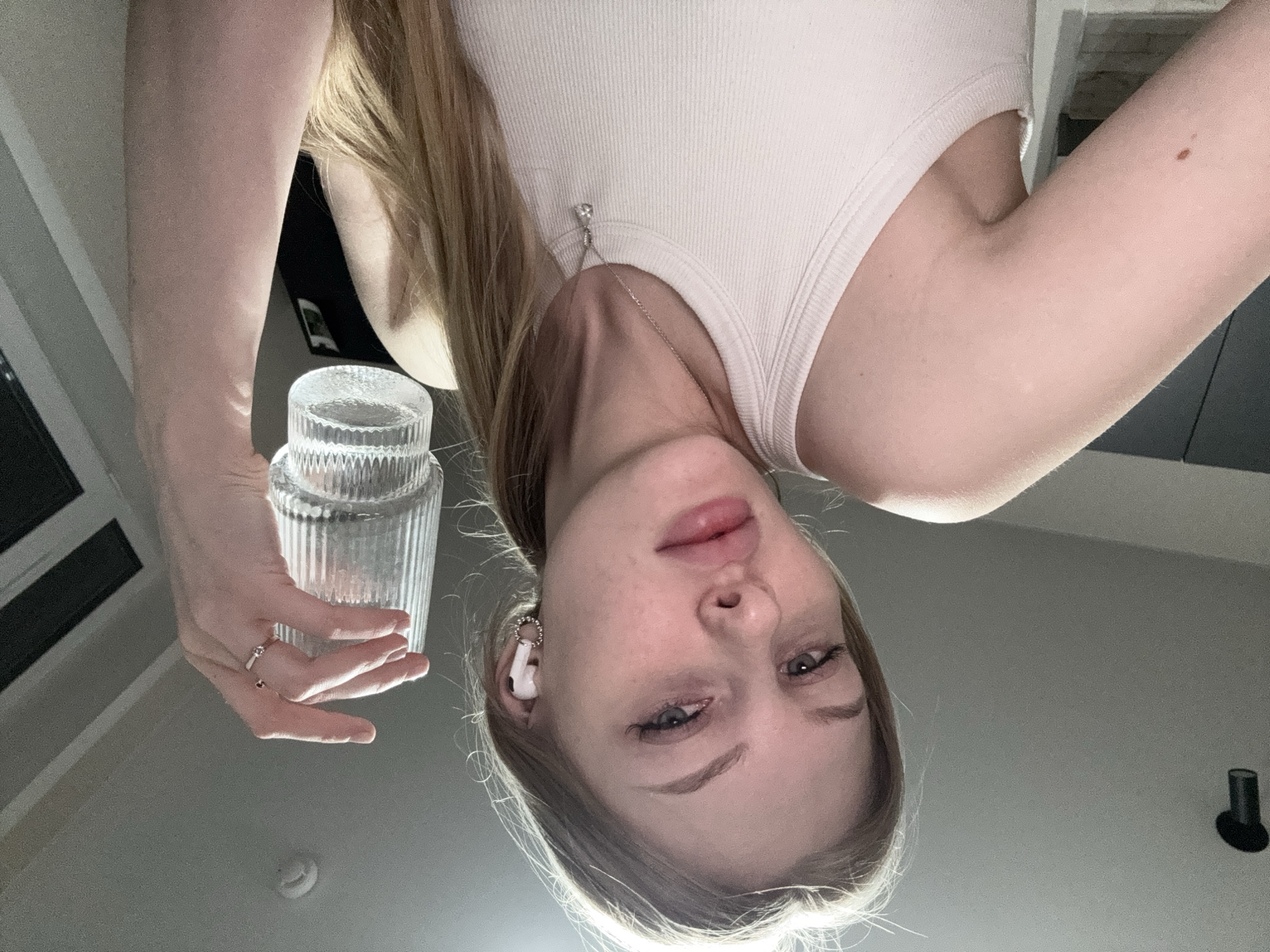 An inverted full-frame, low-angle photograph features a young woman looking directly at the camera. Her fair-skinned face is central, with light-colored hair framing it. She has blue-grey eyes, slightly parted lips, and her expression is neutral. A white earbud, adorned with a decorative silver ring, is visible in her left ear. She wears a plain white ribbed tank top and a silver chain necklace with a small pendant.
Her arms extend downwards from the top of the frame. Her right hand, wearing a silver-colored ring with a stone on the ring finger, holds a clear, textured, ribbed glass or plastic container. Her upper right arm shows a small brown mole. The background is a plain, light-colored ceiling or wall, consistent with an indoor setting. Faint dark lines and a small circular object, possibly a smoke detector, are visible on the ceiling. A dark, cylindrical fixture, possibly a light or camera, is visible in the upper right background. The image is taken from a perspective looking upwards at the woman, likely from a surface she is looking down upon. This scene is located in Perm, Russia. No visible text is present.