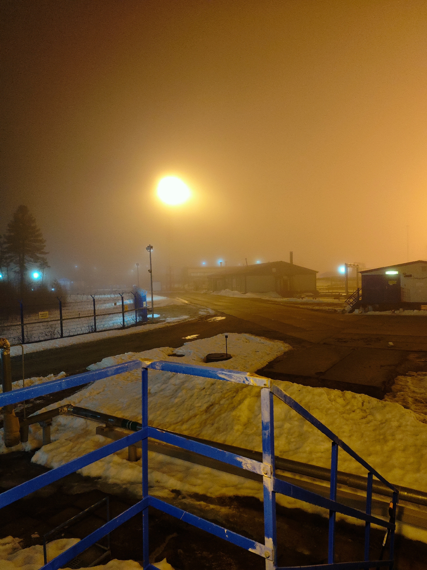 The image depicts a foggy night scene in what appears to be an industrial or utility area. In the foreground is a blue metal railing with snow underneath. Beyond the railing, a road or pathway leads towards buildings illuminated by several lights. A tall street light is visible in the center of the frame. A fence with what looks like barbed wire runs along the left side of the road. The sky is overcast with a yellowish or orange tint. The location is Unknown, Russia. The image contains no people, focusing instead on the infrastructure and environment.