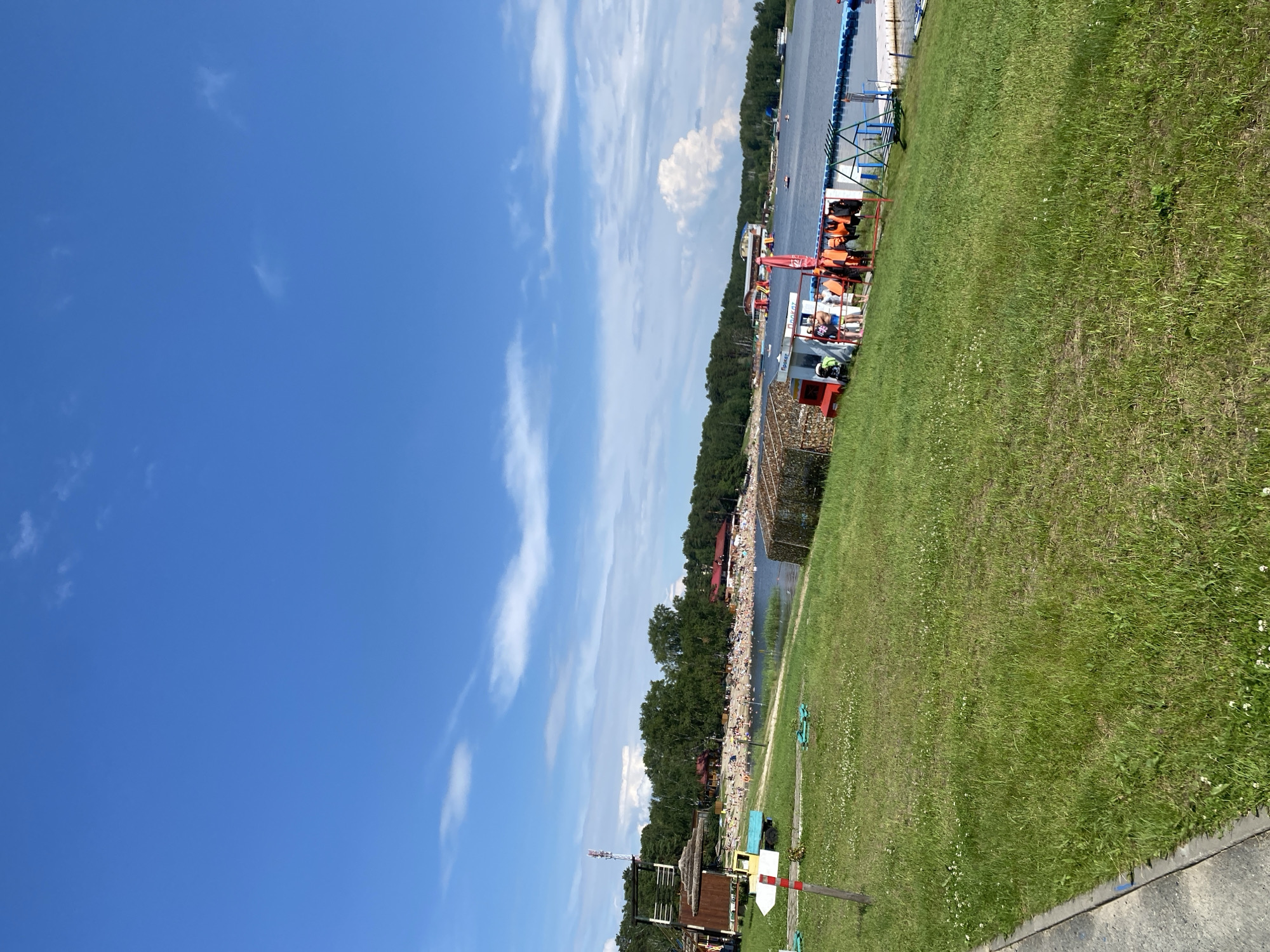 This is a full, eye-level, outdoor, vertical shot of a crowded beach on a sunny day. The sky is bright blue with scattered white clouds. In the foreground, a grassy slope leads down to the beach area. A white directional sign with red stripes points to the left. A small wooden structure with a thatched roof is visible on the left. Further down the beach, many people are sunbathing and swimming. The beach is lined with trees and several buildings with red roofs. A body of water, likely a lake, stretches across the middle ground and background, with a blue floating platform and some boats visible. On the right side of the image, a small red and white ticket booth with life vests hanging from a rack is situated near the water. The scene appears to be a recreational area.