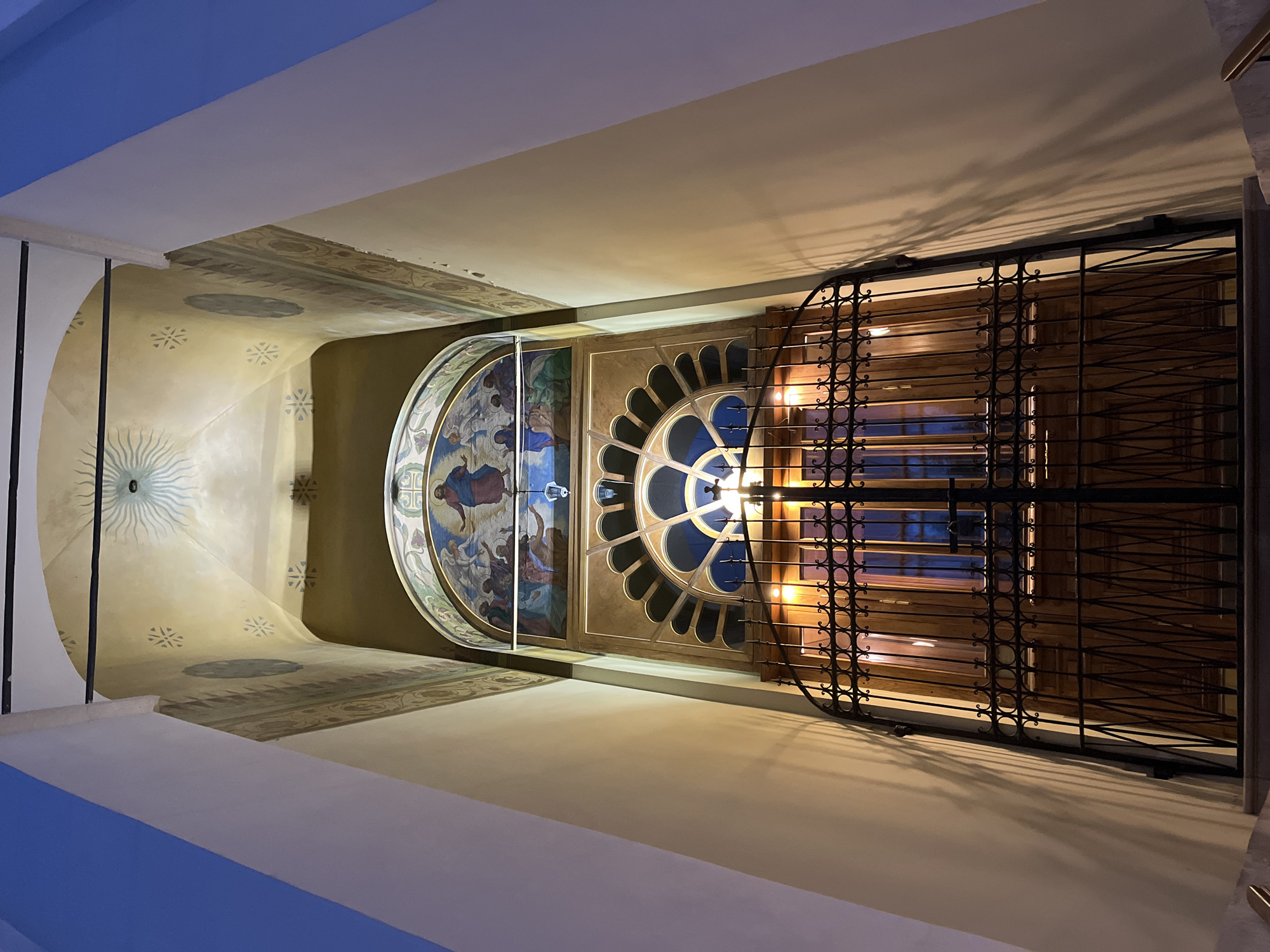 The image is an upward-facing view of an interior architectural space. A section of a domed ceiling is visible, adorned with a painted fresco depicting figures in classical style. To the right of the fresco is a circular window with radial mullions, beyond which a barred gate obscures the view. The surrounding walls are painted in a neutral tone and feature decorative patterns near the ceiling line. The lighting casts shadows on the walls. The location and city are not identified in the given image.