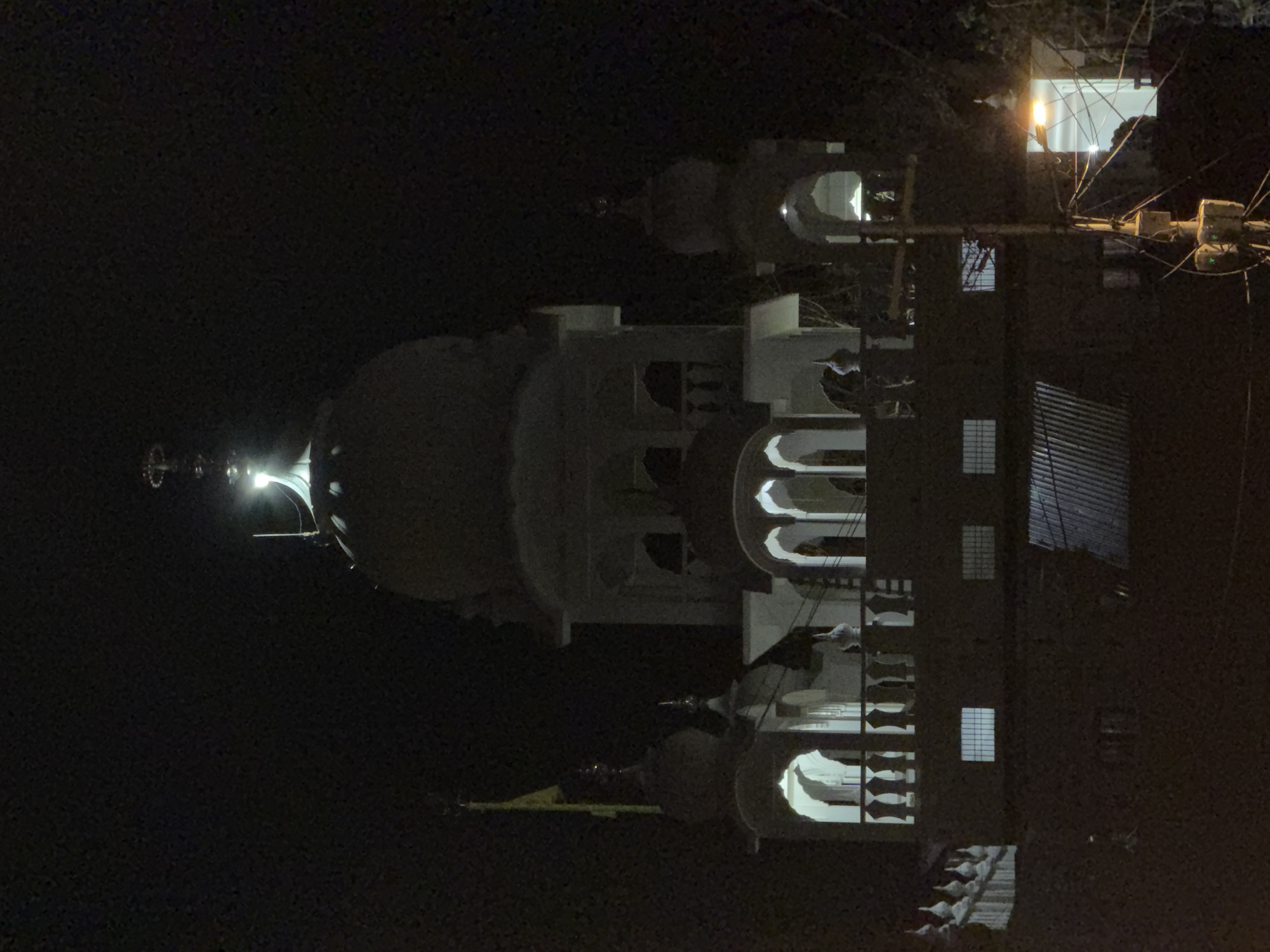 The image depicts a Sikh Gurdwara in Leh, India, illuminated at night. The central structure features a prominent white dome topped with a finial, flanked by smaller domed structures and minarets. Arched windows with ornamental railings are visible on the upper floors, with interior lights casting a warm glow. A yellow flag flies from a pole on the left side of the building. The foreground shows the lower section of the Gurdwara, with a corrugated metal roof and several square windows. A streetlight casts a bright, yellow-orange light on the right side of the image, illuminating a pole with numerous wires. The overall scene is dark, with the architecture of the Gurdwara being the primary illuminated subject.