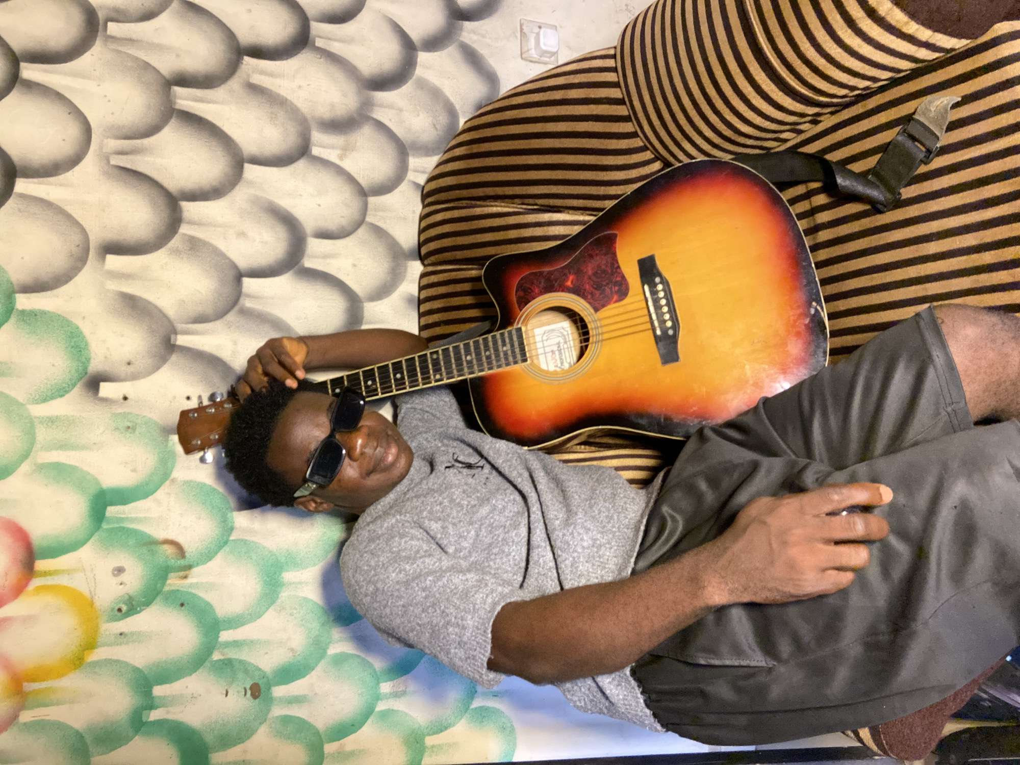 An overhead shot shows a man relaxing indoors with an acoustic guitar. The man, with dark skin, is lying on his back, facing slightly towards the camera. He wears dark sunglasses, a light grey short-sleeved sweater, and dark grey shorts. His left arm is bent, resting near his head, while his right hand rests on his shorts.
An acoustic guitar with a sunburst orange and brown finish lies on his torso and to his side. His left hand is positioned near the headstock and fretboard of the guitar. The guitar has six strings, a pickguard, and a label visible through its soundhole, though the text is unreadable. A black strap is attached to the guitar and draped over the striped furniture next to him.
The man is positioned on a surface next to a piece of furniture upholstered with brown and black horizontal stripes. Behind him is a wall with a distinctive pattern of large, overlapping, scale-like or bubble-like shapes. These shapes are predominantly grey and white, with some green, yellow, and red hues visible in the bottom-left corner of the frame. A white wall socket is visible on the wall above the striped furniture. The scene appears to be an indoor, relaxed setting, consistent with a home environment in Mararaba, Nigeria.
