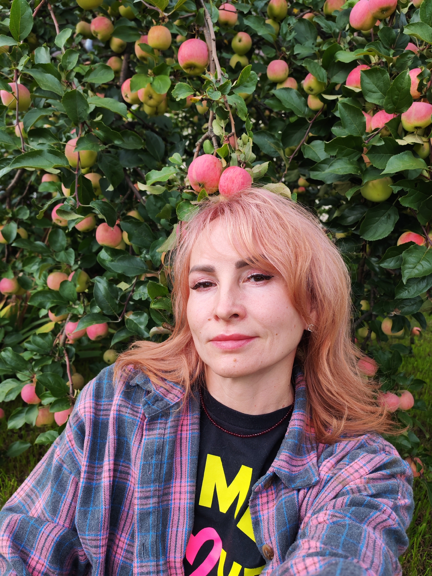 A medium shot captures a woman with light pink hair and makeup, wearing a plaid shirt over a black t-shirt with yellow and pink graphic print. She is positioned in front of a tree laden with ripe red and green apples. The background consists of lush green foliage and grass. The image appears to have been taken outdoors, possibly in a garden or orchard. No specific location in Chaikovsky, Russia is identifiable from the image itself.