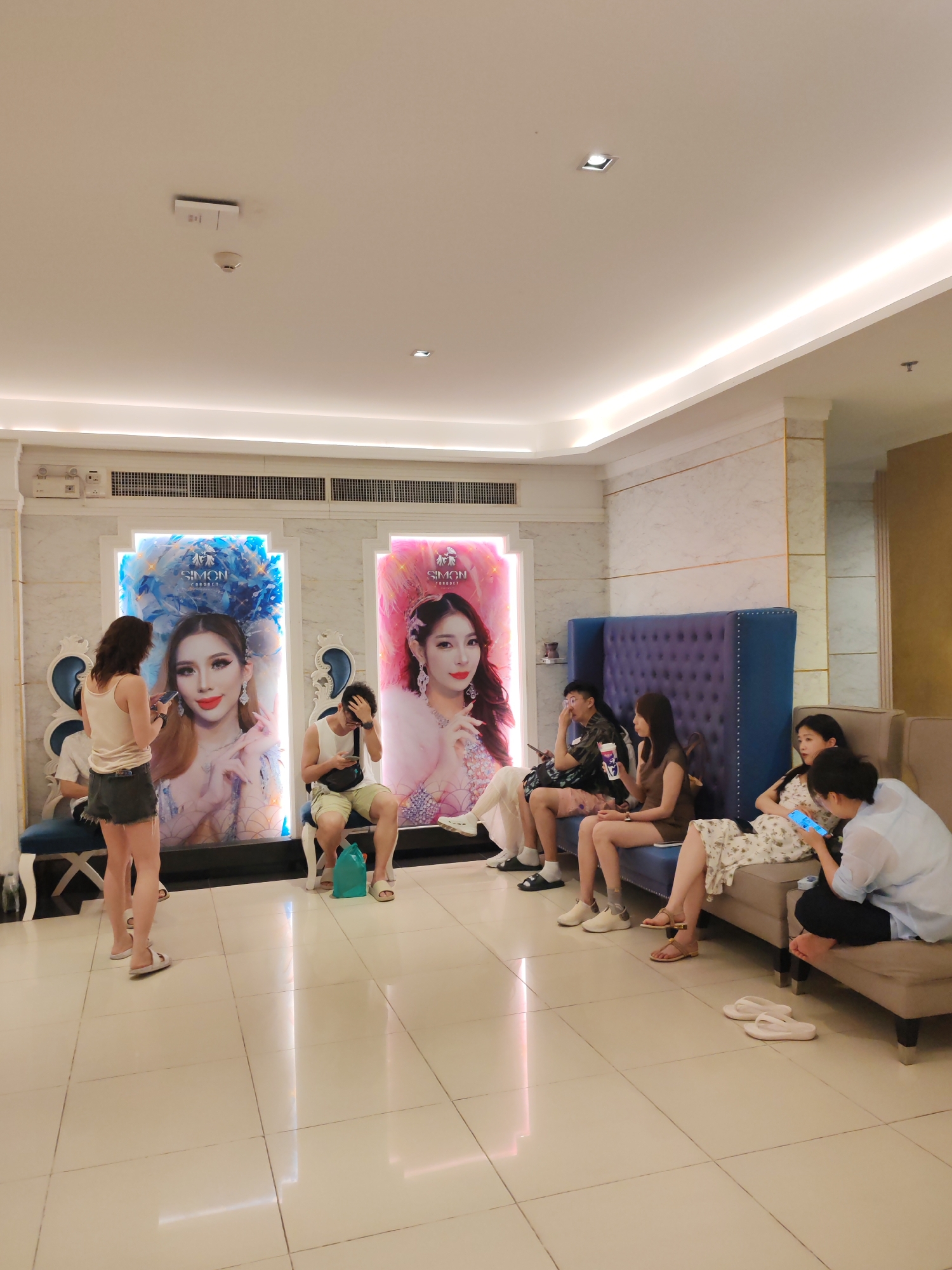 The image depicts an indoor waiting area, likely in a business such as a cosmetic surgery center, based on the presence of large posters of female models with the name "Simon" prominently displayed. Several people are present. Near the posters, two individuals stand; one appears to be taking a picture with a smartphone, while another is sitting. A group of people is seated on a long sofa and a blue high-backed chair, with some using their phones. The environment is well-lit with tiled flooring and modern ceiling lights. The overall impression is of a modern, upscale waiting room. The location is assumed to be in Patong, Thailand based on the prompt.