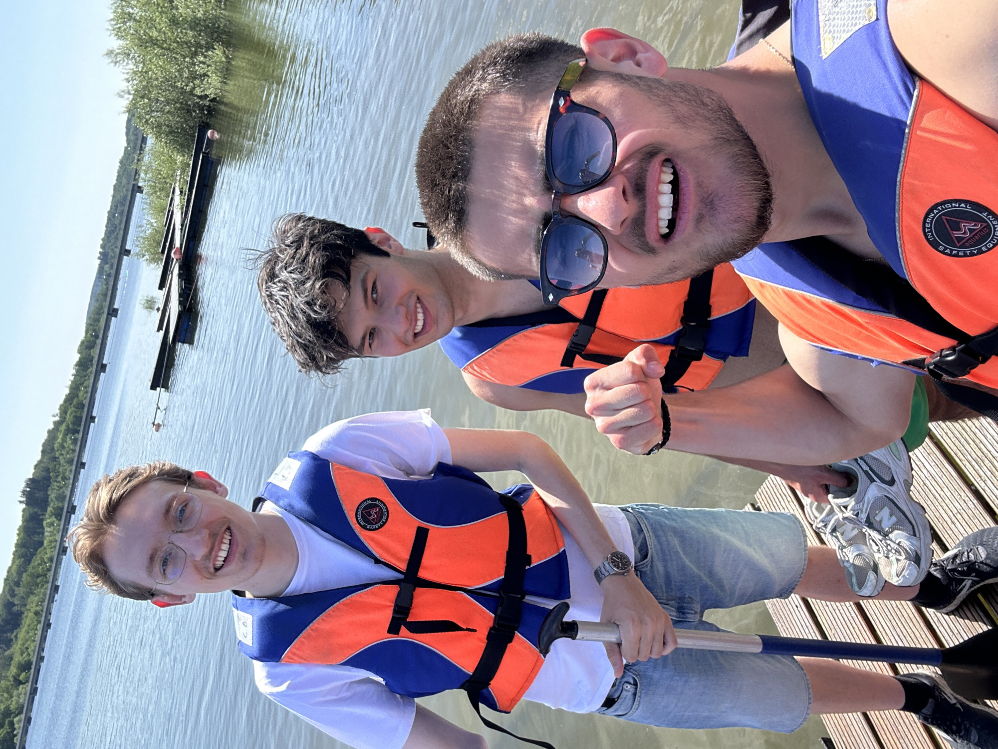 Three young men are on a wooden dock at Möhnesee, Germany. All three are wearing orange and blue life vests over white t-shirts. The man closest to the camera has glasses and is holding a paddle. The man next to him is smiling. The third man is wearing sunglasses and is also smiling. The water and trees can be seen in the background.