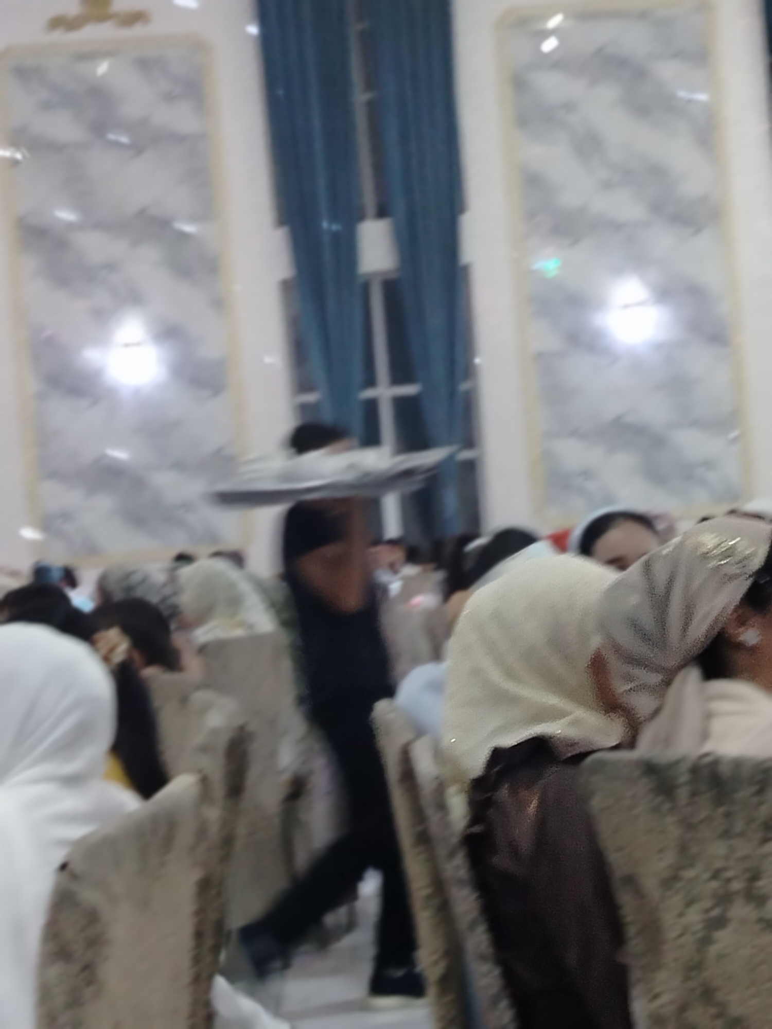 In Ақсуабат, Kazakhstan, a person in a black shirt is carrying a large platter over their head, walking through a gathering. Several people are seated at tables, some are wearing headscarves. The environment appears to be an indoor event space with decorative panels on the walls and blue curtains.