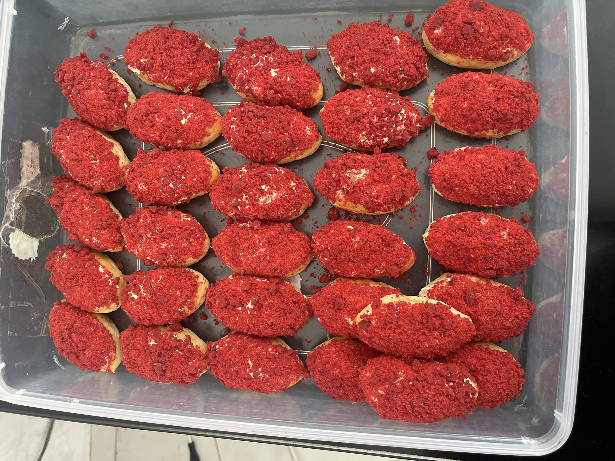 A clear plastic rectangular container holds multiple rows of small, oval-shaped pastries. Each pastry has a light-colored base, possibly cake or dough, topped with a white filling. The pastries are generously coated with a vibrant red, crumbly topping. The container is filled with these pastries arranged closely together, with some resting on a wire rack within the container. The background is dark and out of focus.