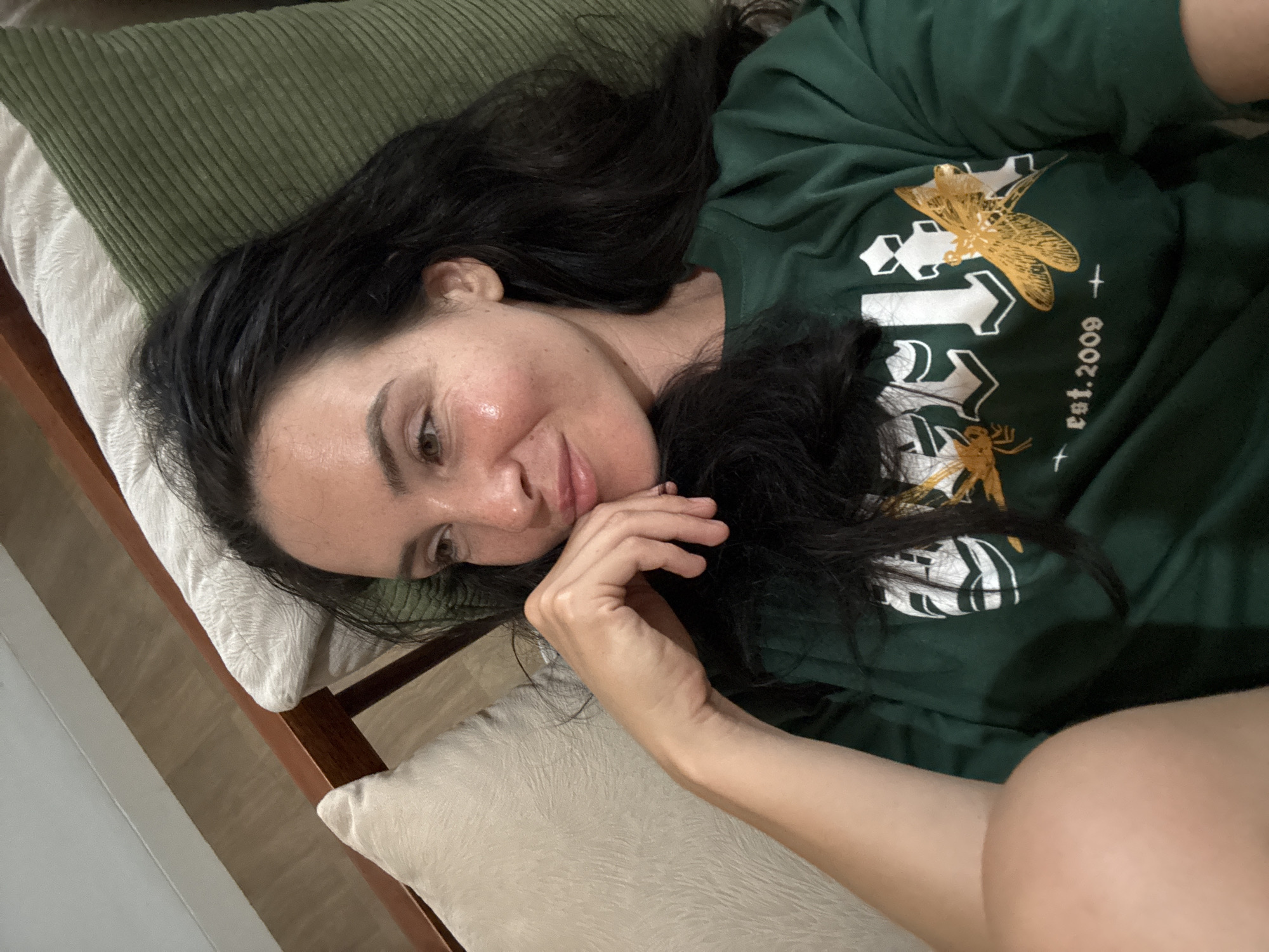 A woman with dark hair is lying on a couch, with her head resting on a green corduroy pillow. She is wearing a dark green t-shirt with a graphic design that includes the text "est. 2009" and two stylized insects. Her left hand is supporting her chin as she looks off to the side with a slight smile. The couch has a wooden frame and other pillows visible. The lighting suggests an indoor setting, possibly in North Kuta, Indonesia.