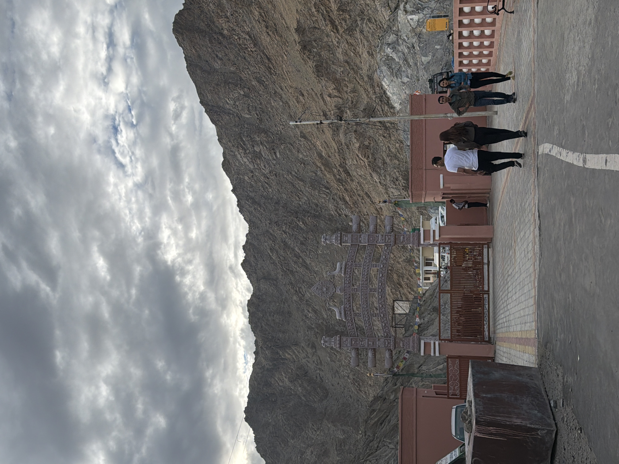 The image depicts an entrance gate in Leh, India, under a cloudy sky. The gate is ornate, with decorative carvings and Buddhist motifs. Behind the gate, a road leads to buildings, and a steep, rocky mountain dominates the background. Four individuals, two couples, are walking away from the camera on a paved pathway leading to the gate. The couple in the foreground consists of a man in a white t-shirt and black pants and a woman in a dark jacket and black pants. The couple behind them consists of a man in a green jacket and jeans and a woman in a blue jacket and black pants. A utility pole with wires stands to the right of the path. The area is paved with interlocking bricks and has a low, decorative pink wall with white circular elements to the right.
