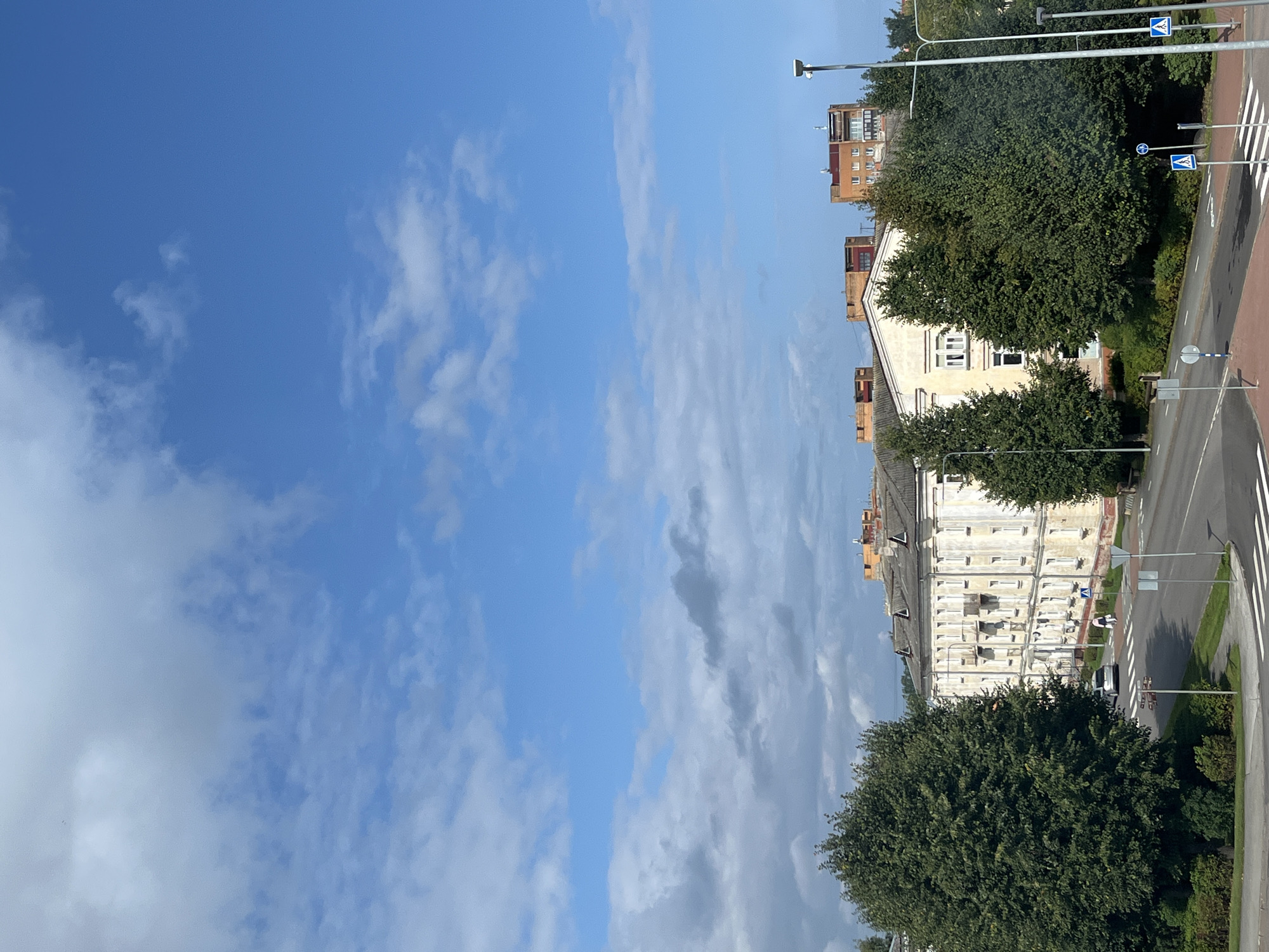 This image captures a street view in Kohtla-Järve linn, Estonia. A multi-story apartment building with a light-colored facade and a grey roof dominates the middle ground, situated alongside a paved road. To the left of the building, a large, dense green tree stands prominently. Across the street, more trees and smaller buildings are visible, including some taller, orange-hued structures in the background. The sky is a bright blue with scattered white clouds. A crosswalk is marked on the road in the foreground, and traffic signs are present. A single person is visible walking across the crosswalk.